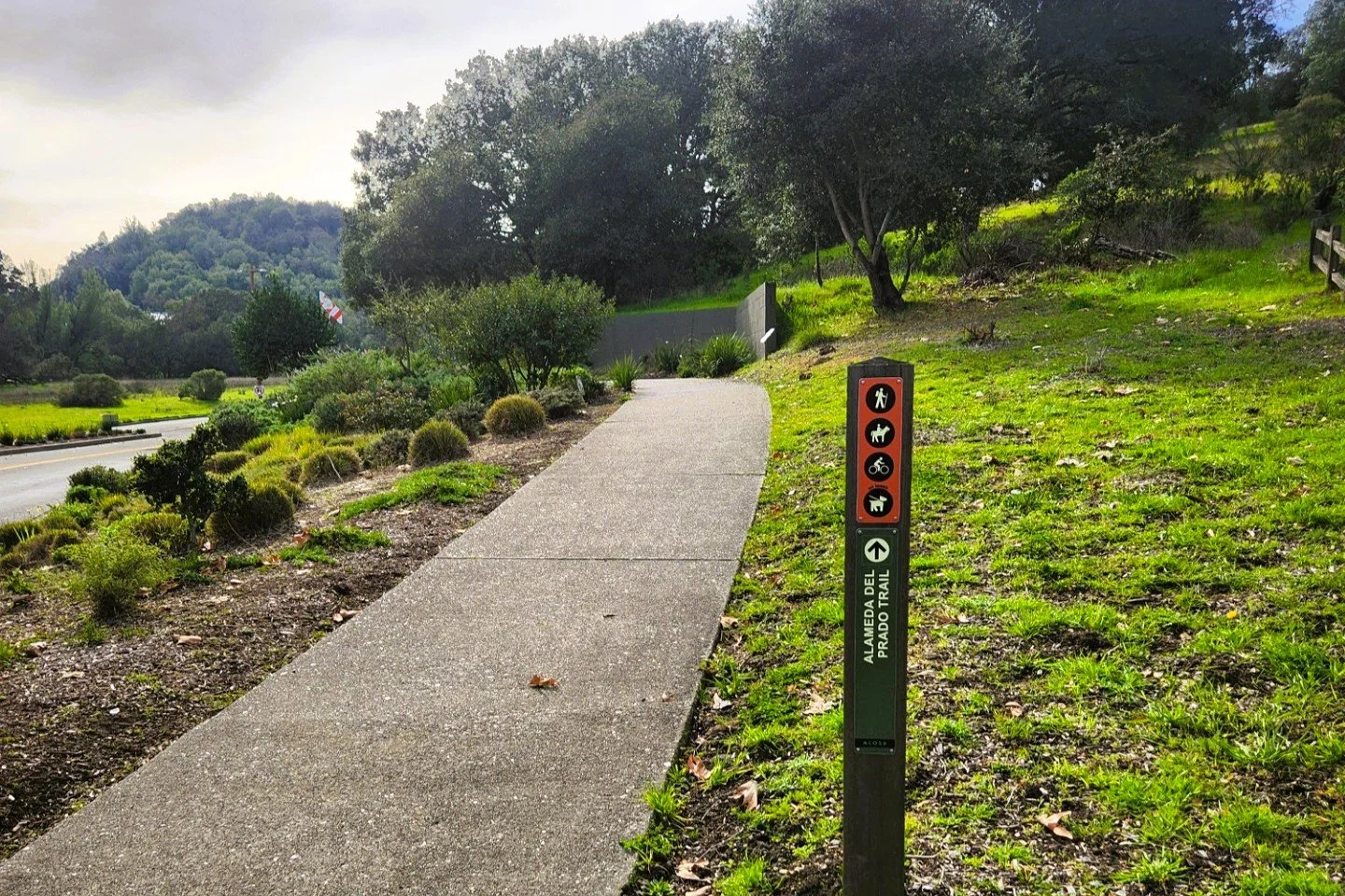 A sidewalk alongside a green grassy area with trees and bushes, featuring a trail marker sign that indicates the trail is for pedestrians, dogs, bicycles, and strollers, located in Alameda del Prado.