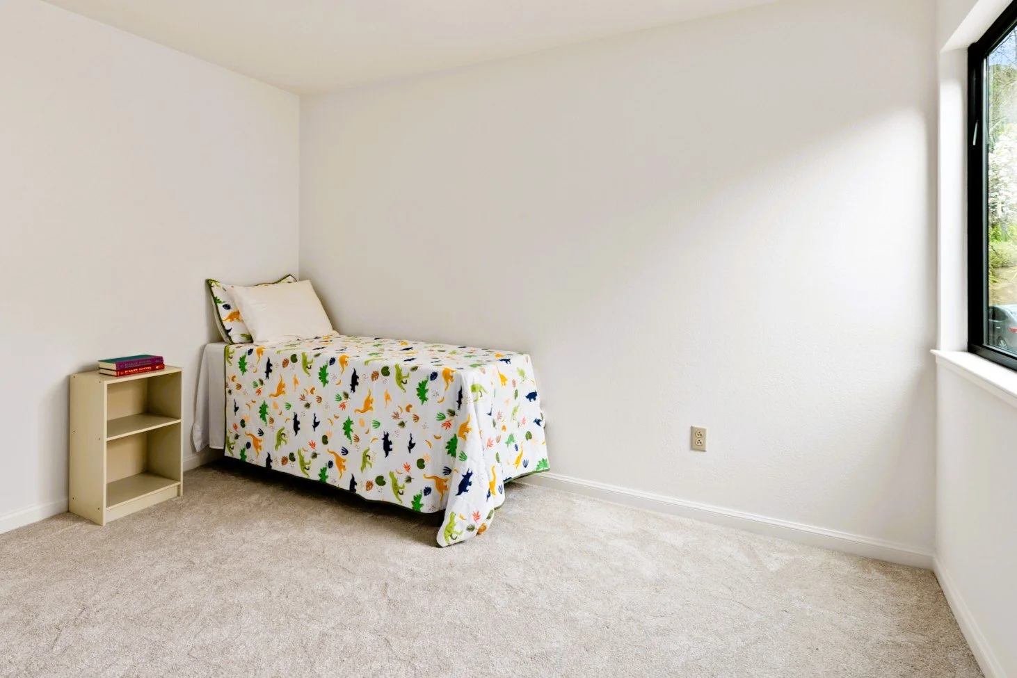 A bedroom with a single bed covered in a colorful dinosaur-themed bedspread, a small beige bookshelf with a few books, and a window letting in natural light.