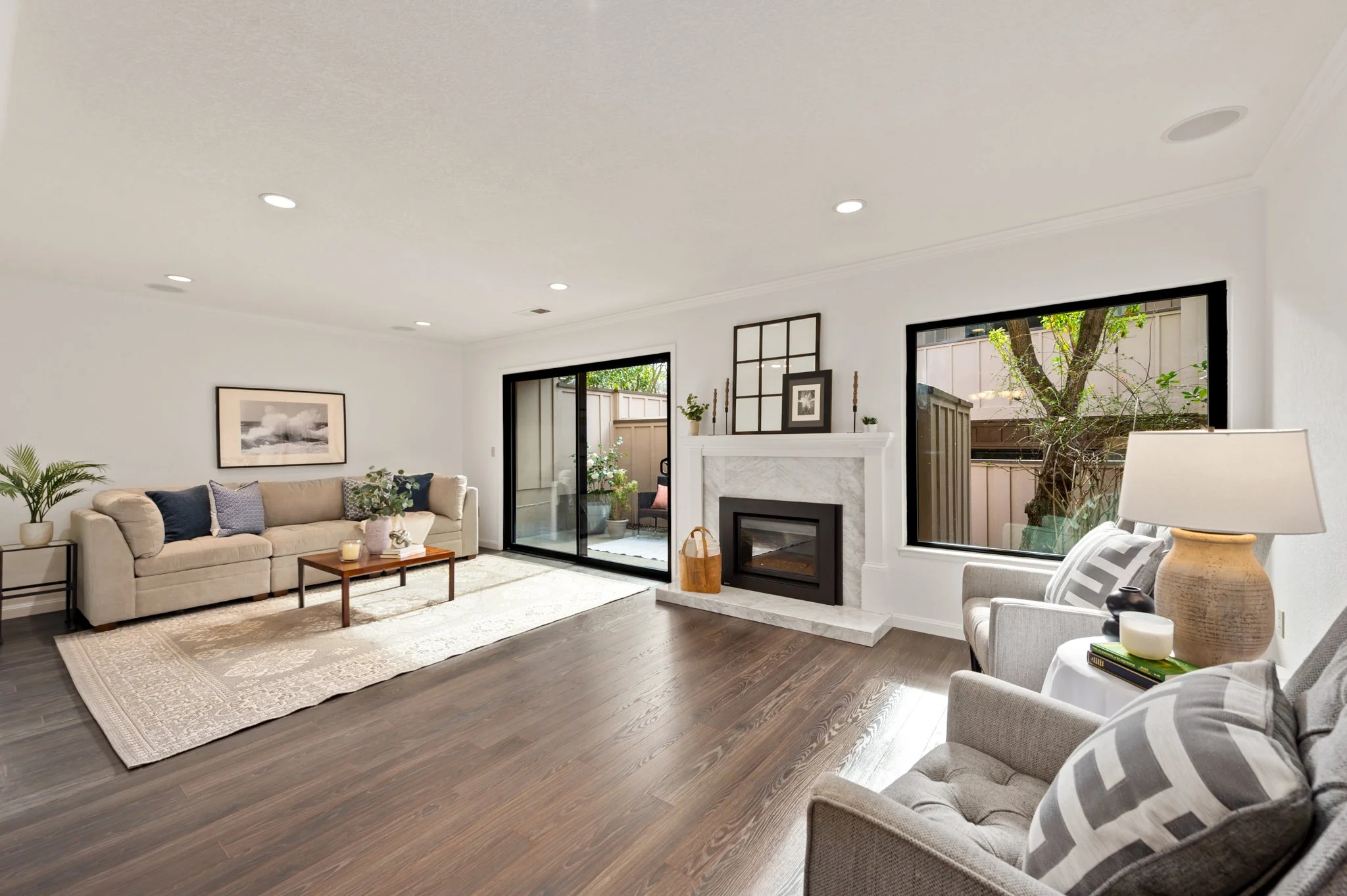 Living room with beige sofa, gray armchair, fireplace, large windows, wooden flooring, potted plants, and modern decor.