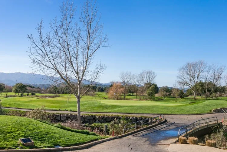 A golf course with green fairways, leafless trees, a small bridge over a creek, and mountains in the background under a clear blue sky.