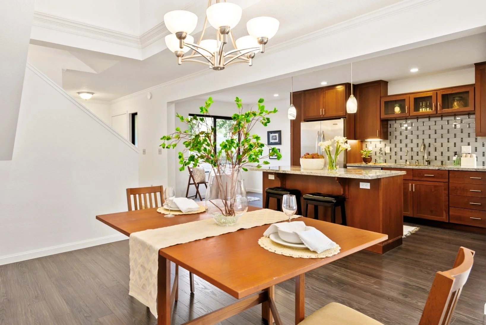 Open concept kitchen and dining area with a wooden table set with plates, napkins, wine glasses, and a vase with green plants. The kitchen features wooden cabinets, a granite countertop, stainless steel refrigerator, pendant lights, and a backsplash.