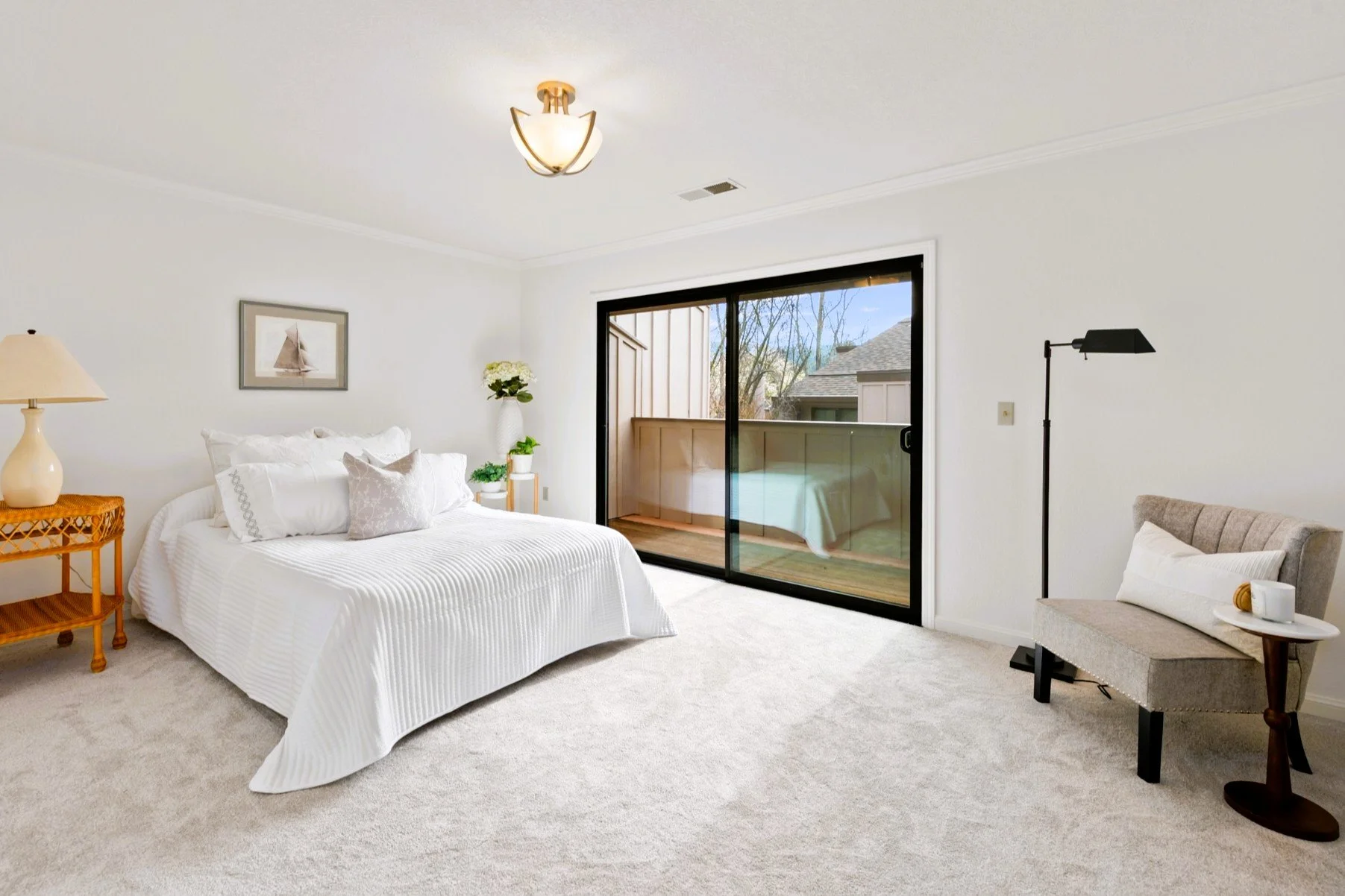 A bedroom with white walls, a bed with white linens, a bedside table with a lamp, a sliding glass door leading to a balcony, a chair with a pillow, and a floor lamp.