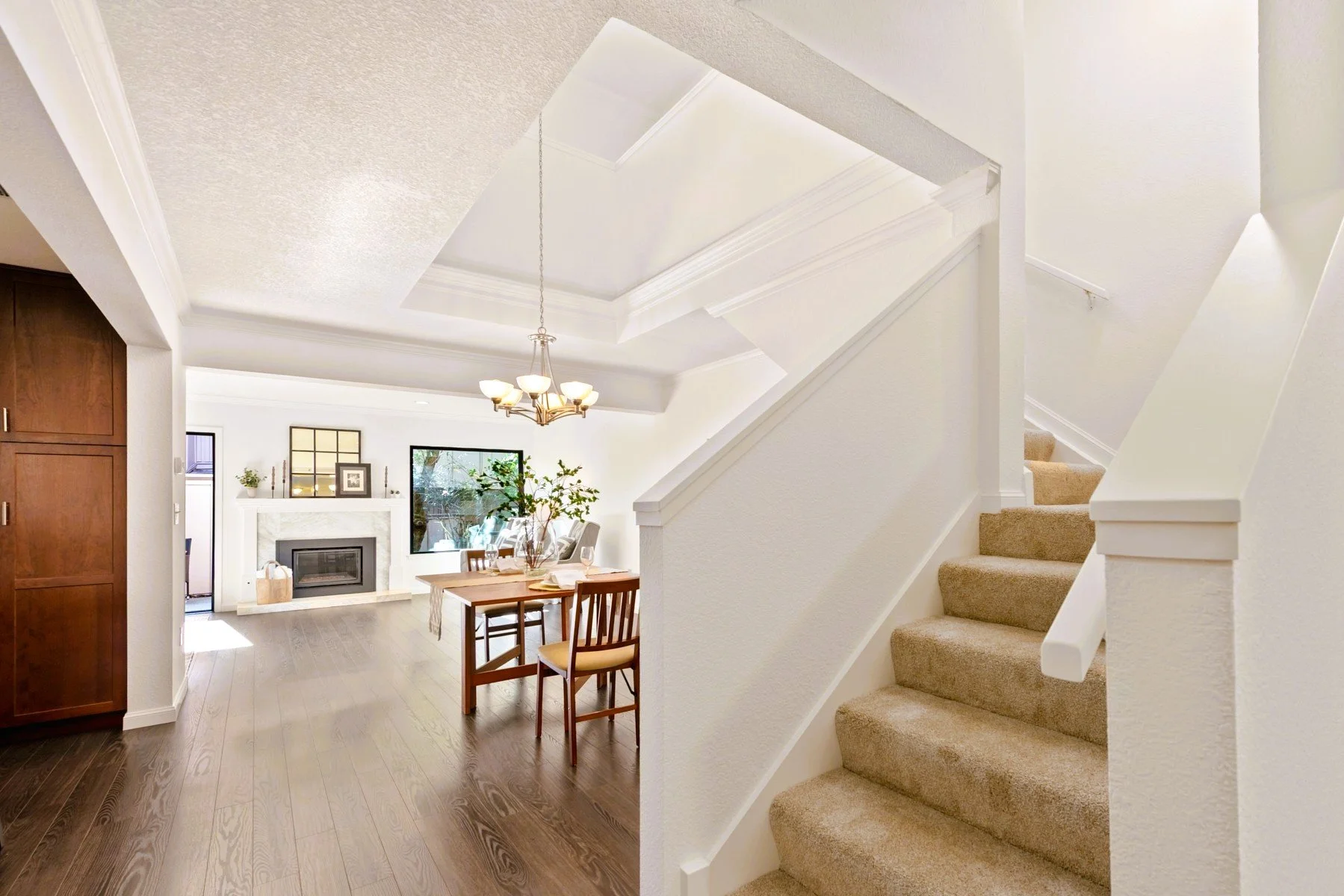 Living room with fireplace, wooden floor, dining table with chairs, staircase with beige carpet, chandelier, and a window showing greenery outside.