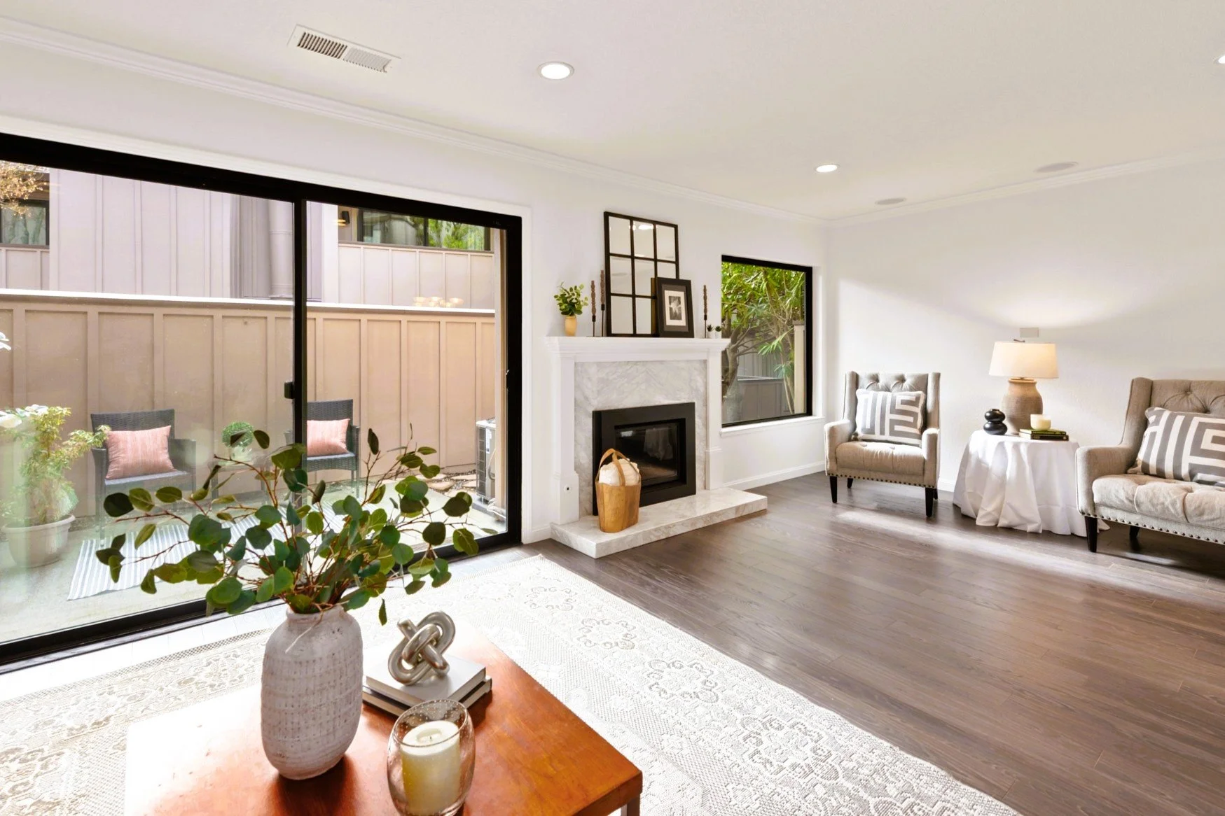 Bright living room with sliding glass door opening to a patio, fireplace with decorative items on the mantel, two armchairs with patterned cushions, and a side table with a lamp.
