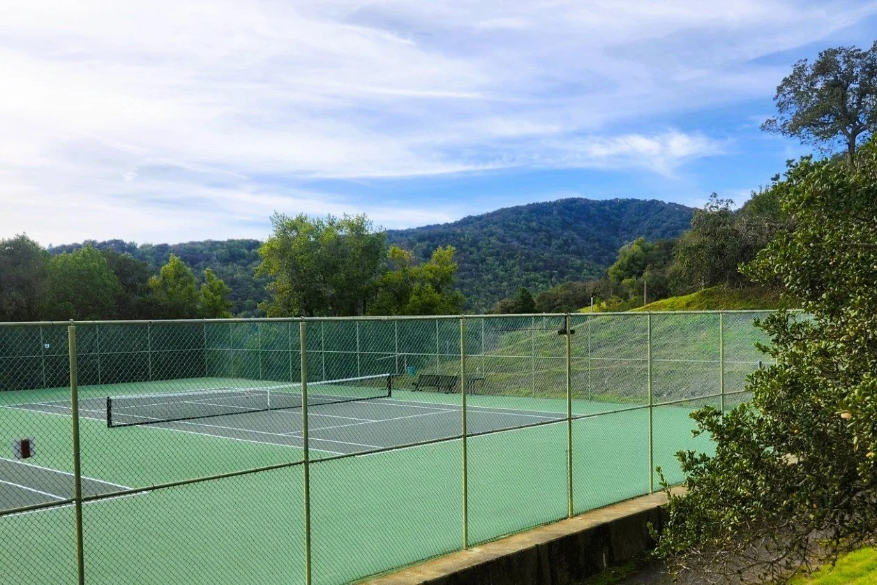 Empty outdoor tennis court with a mountain and trees in the background, partly cloudy sky.