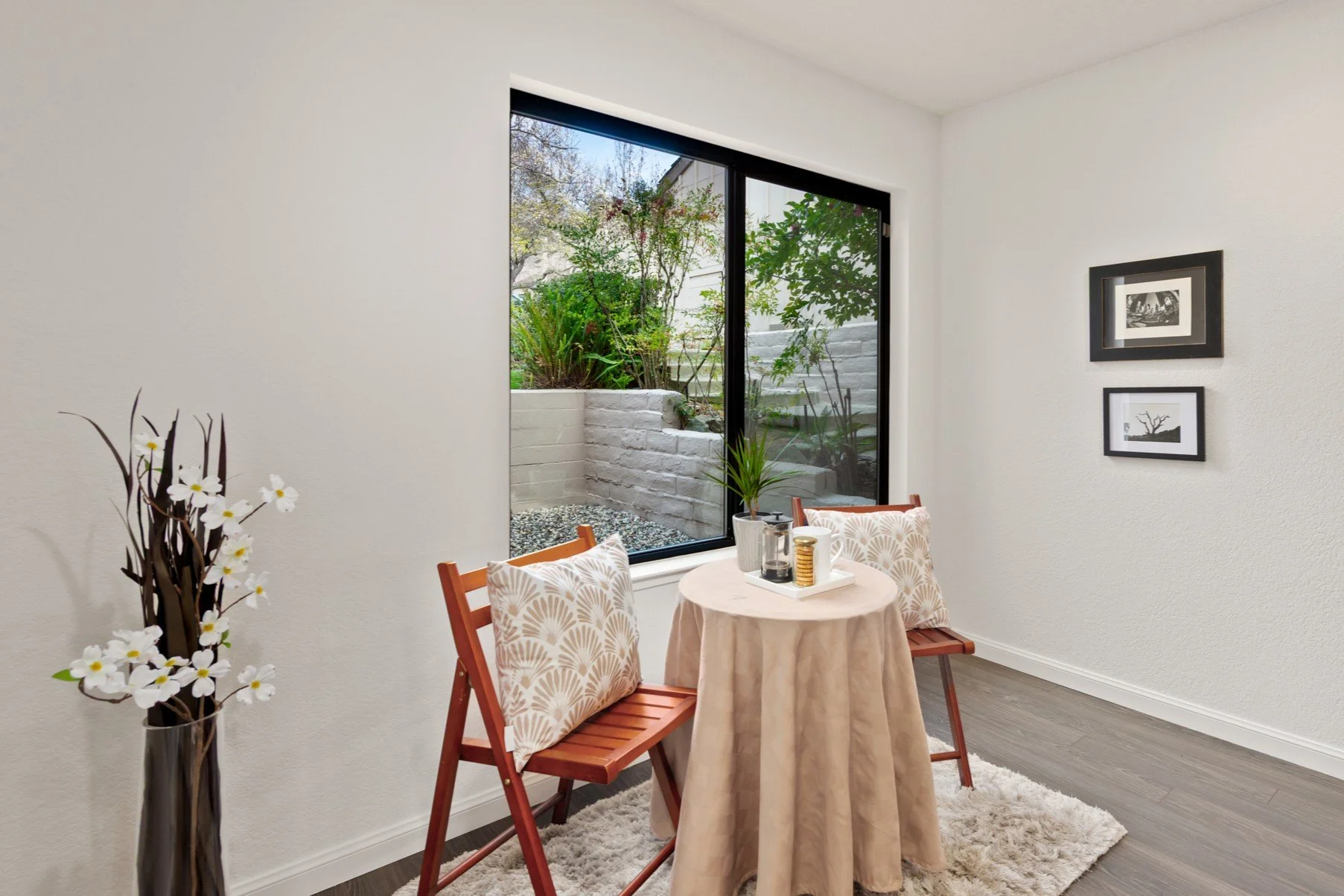 Interior of a bright room with a small round table covered in a beige cloth, two wooden chairs with patterned cushions, a vase with white flowers, and framed black and white artwork on the wall. A large window shows a landscaped outdoor area with pla