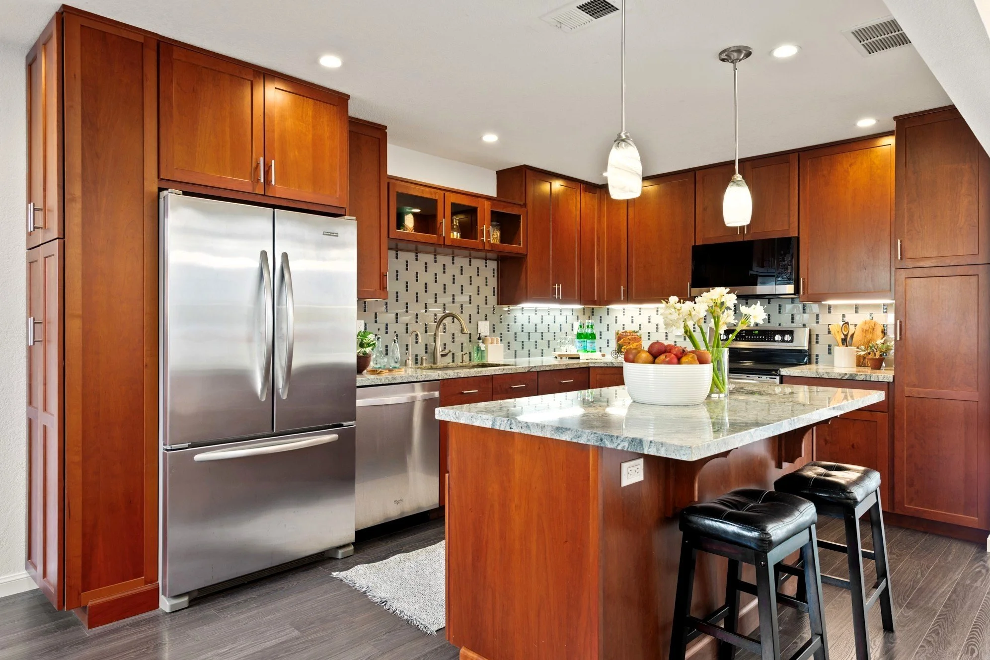 Modern kitchen with wooden cabinets, stainless steel appliances, granite countertops, and a kitchen island with two black stools. Decor includes a fruit bowl and flower vase.