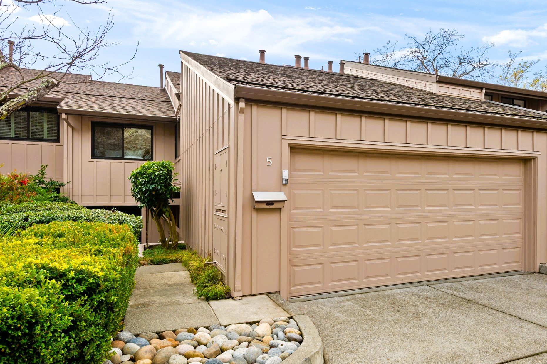 Exterior view of a beige townhouse with a garage, surrounded by green bushes and trees, under a partly cloudy sky.
