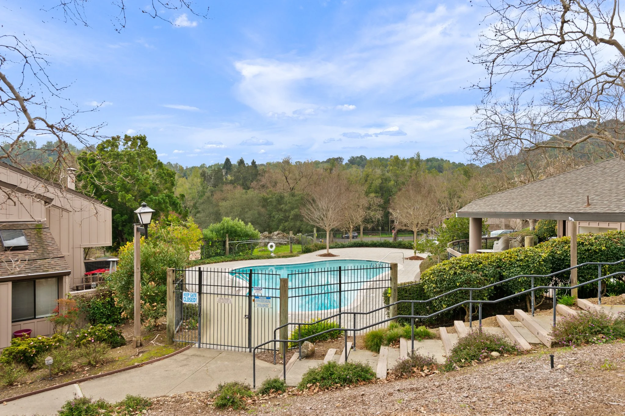 Swimming pool enclosed by black fence, surrounded by trees and shrubbery, with residential buildings nearby, and a backdrop of green hills and a partly cloudy sky.