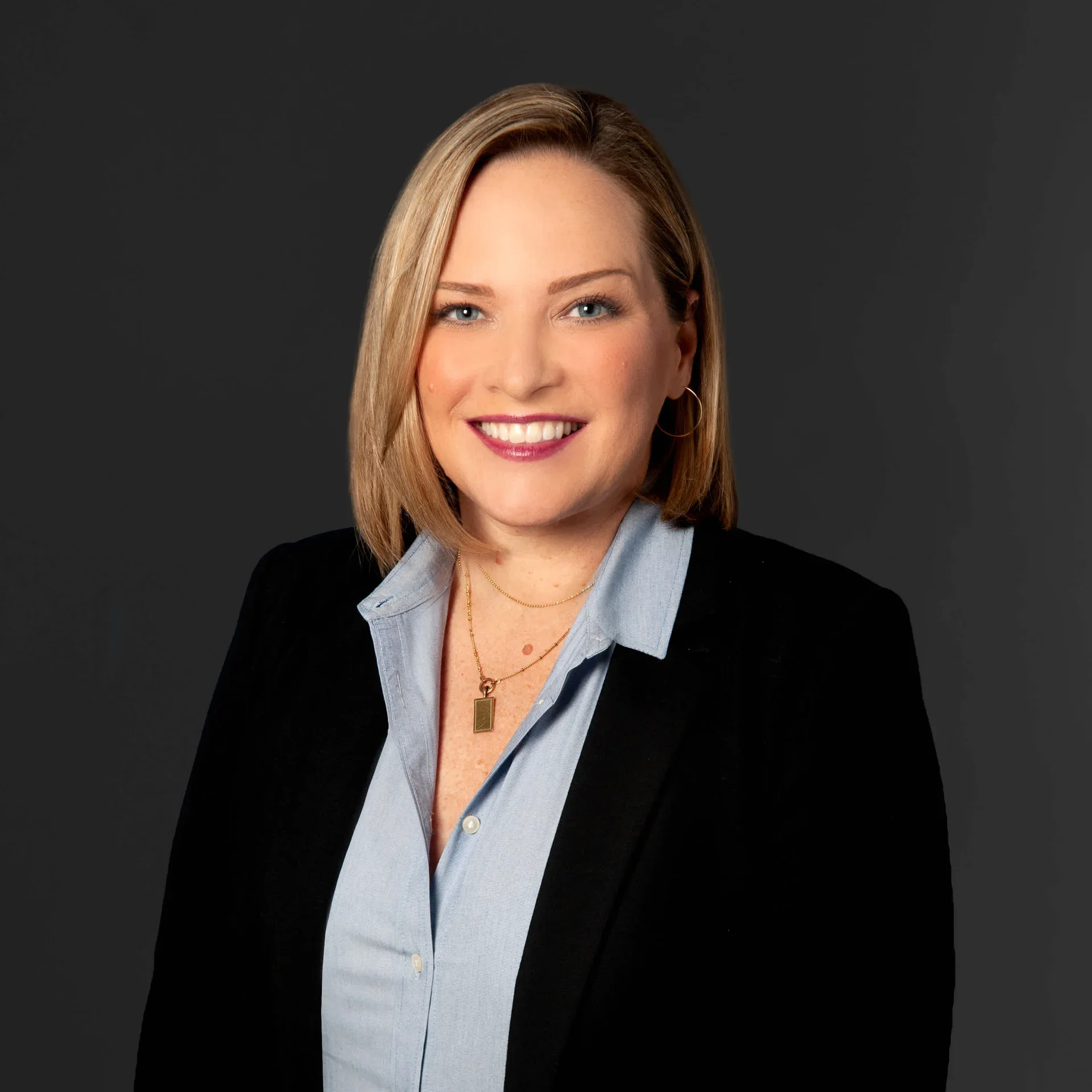 Portrait of a smiling woman with shoulder-length blonde hair, wearing a black blazer over a light blue collared shirt, gold jewelry, and hoop earrings against a dark gray background.