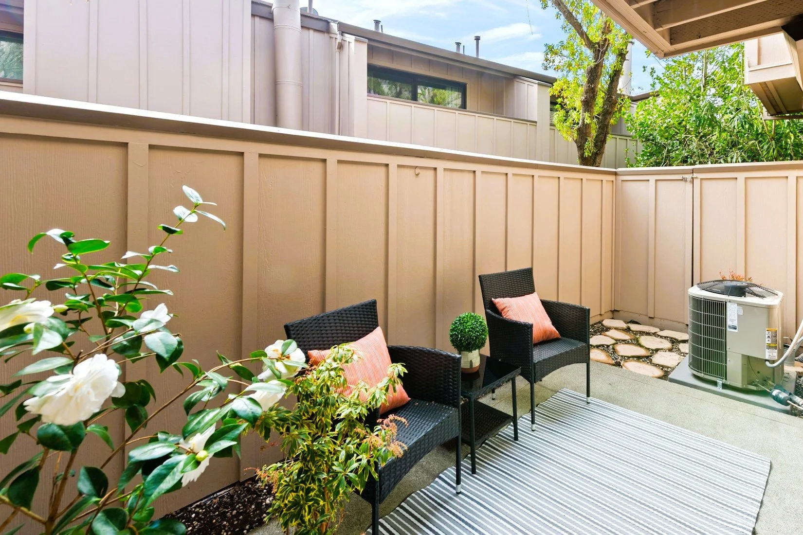 Small outdoor patio with two black wicker chairs with pink cushions, a small table with a green potted plant, a striped rug, plants and bushes near a beige wooden fence, an air conditioning unit, and a tree in the background.