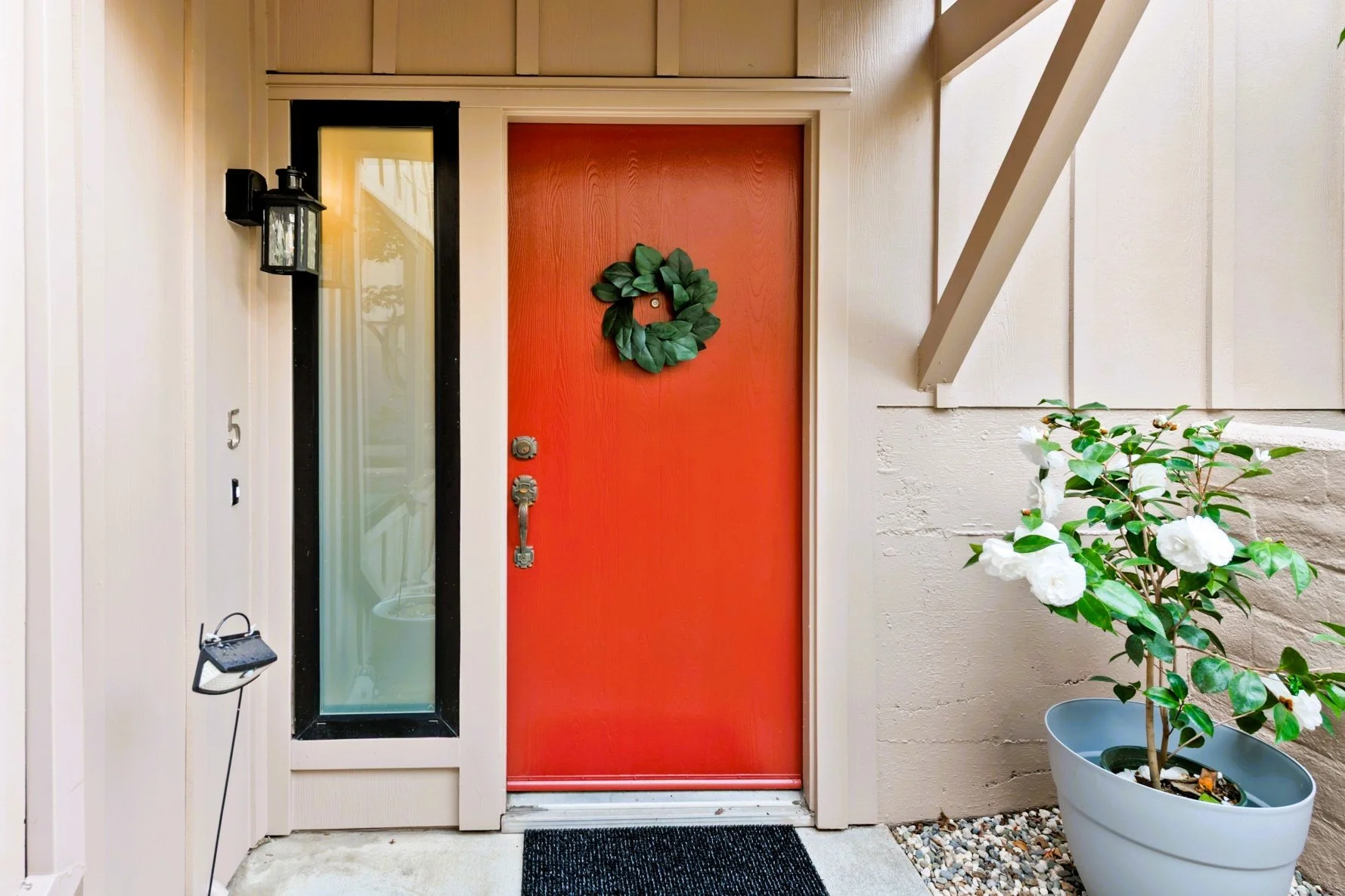 Red front door with a green wreath, next to a glass side panel, with a potted white flower plant on the right.