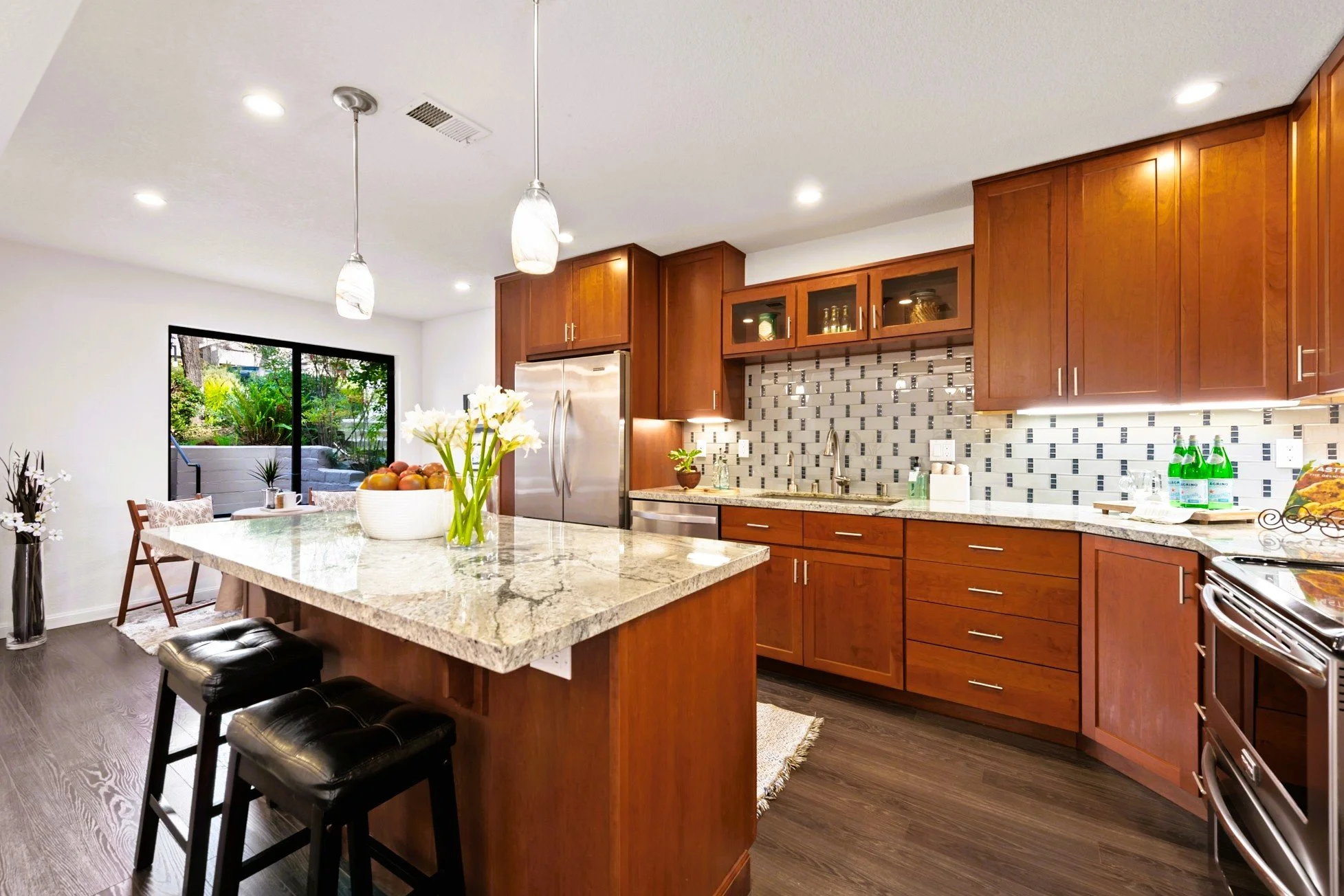 Kitchen with wooden cabinets, granite countertops, mosaic tile backsplash, stainless steel appliances, island with a flower arrangement and fruit, dining area with sliding glass door to outdoor patio, dark wood flooring, recessed lighting, hanging pe