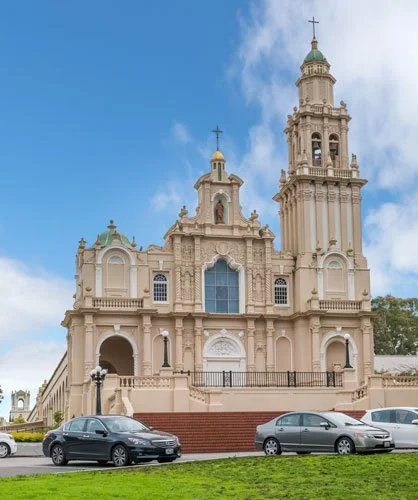 A large, historic church with a tall bell tower, ornate architecture, and multiple arches, located in a city with parked cars in front and green grass.