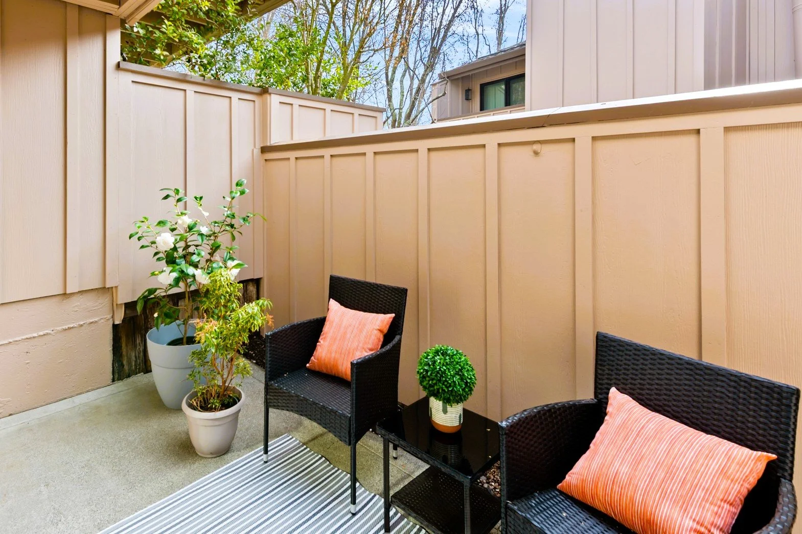 Small outdoor patio with two black wicker chairs, each with orange striped pillows, a small black table with a potted green topiary, and two potted plants against a beige fence. A striped outdoor rug is on the concrete floor.