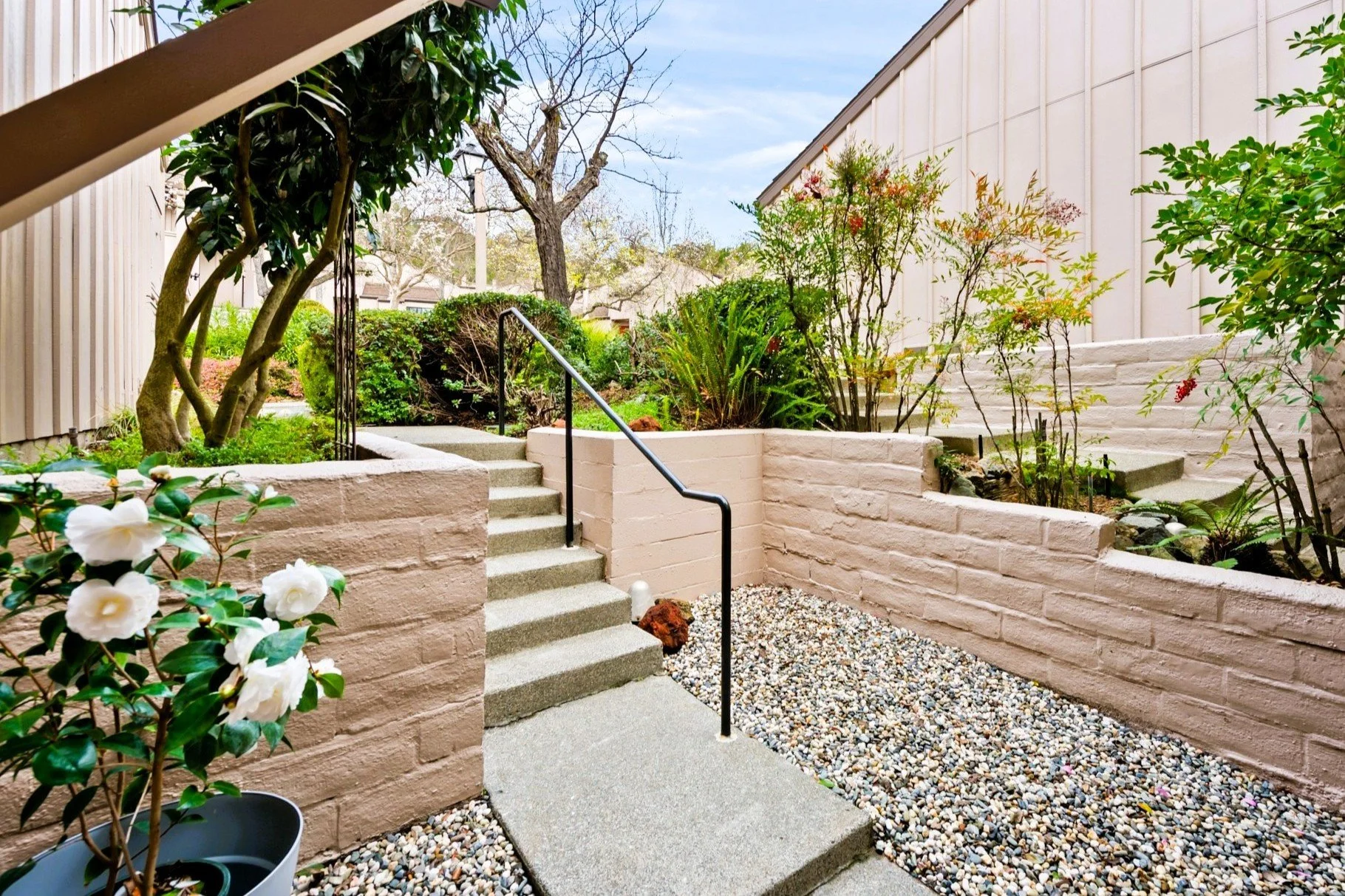 A tiered garden area with beige brick walls, steps, and a black handrail. There are trees, bushes, and flowering plants with a gravel ground cover and a concrete pathway.