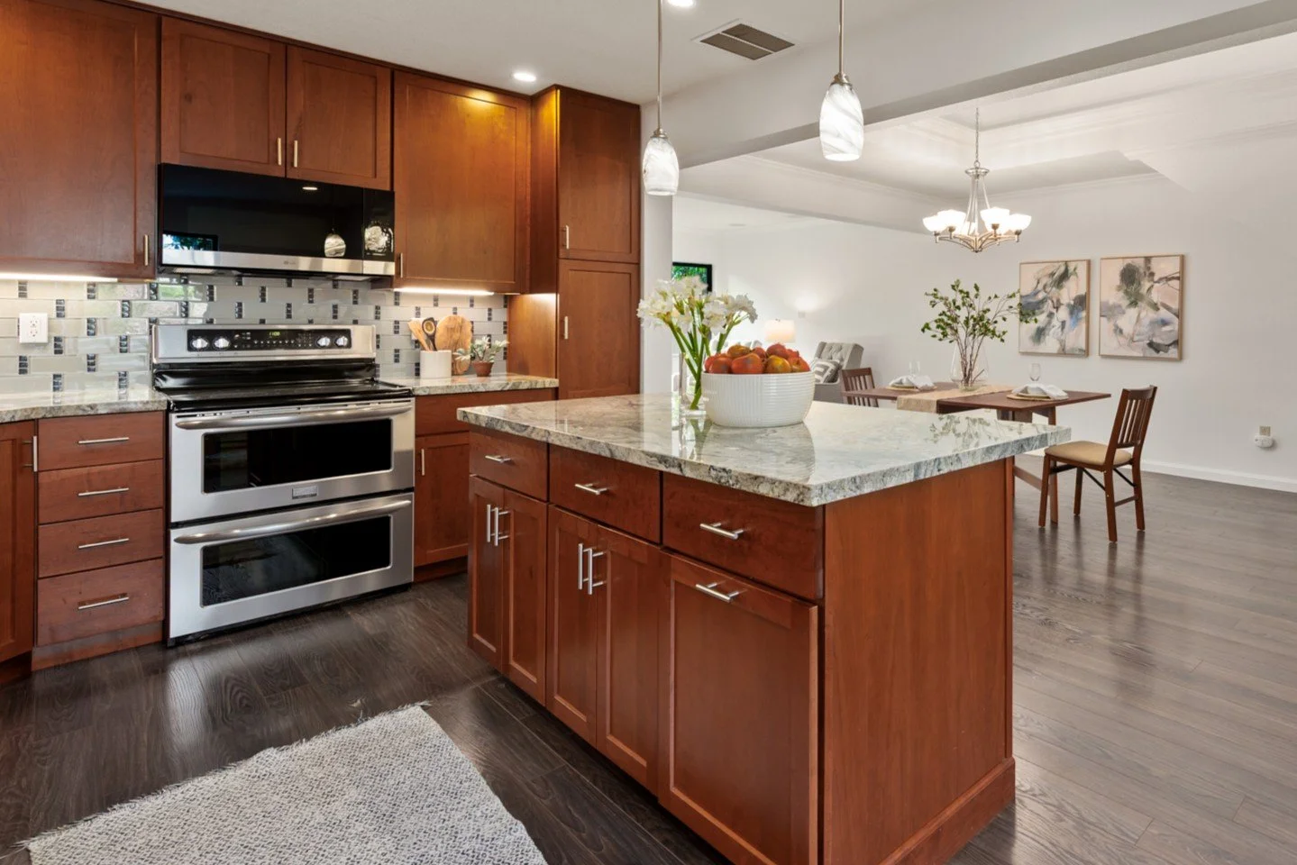 Modern kitchen with wooden cabinets, stainless steel oven and microwave, granite countertops, and a large island with a bowl of fruit. Adjacent dining area with table, chairs, and wall art.