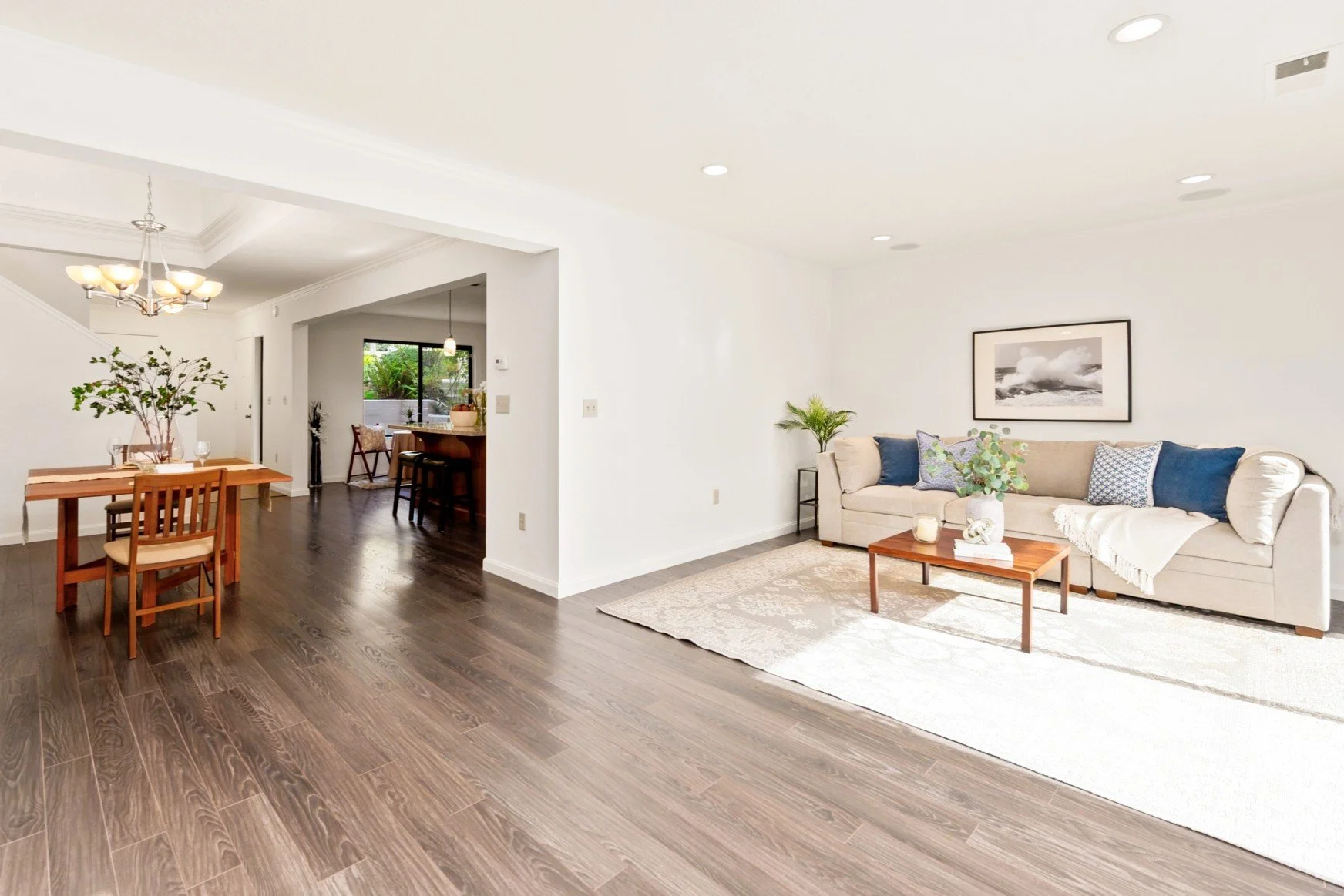 Living room with beige sofa, blue and patterned pillows, wooden coffee table, potted plant, framed black and white art on white wall, and hardwood flooring.
