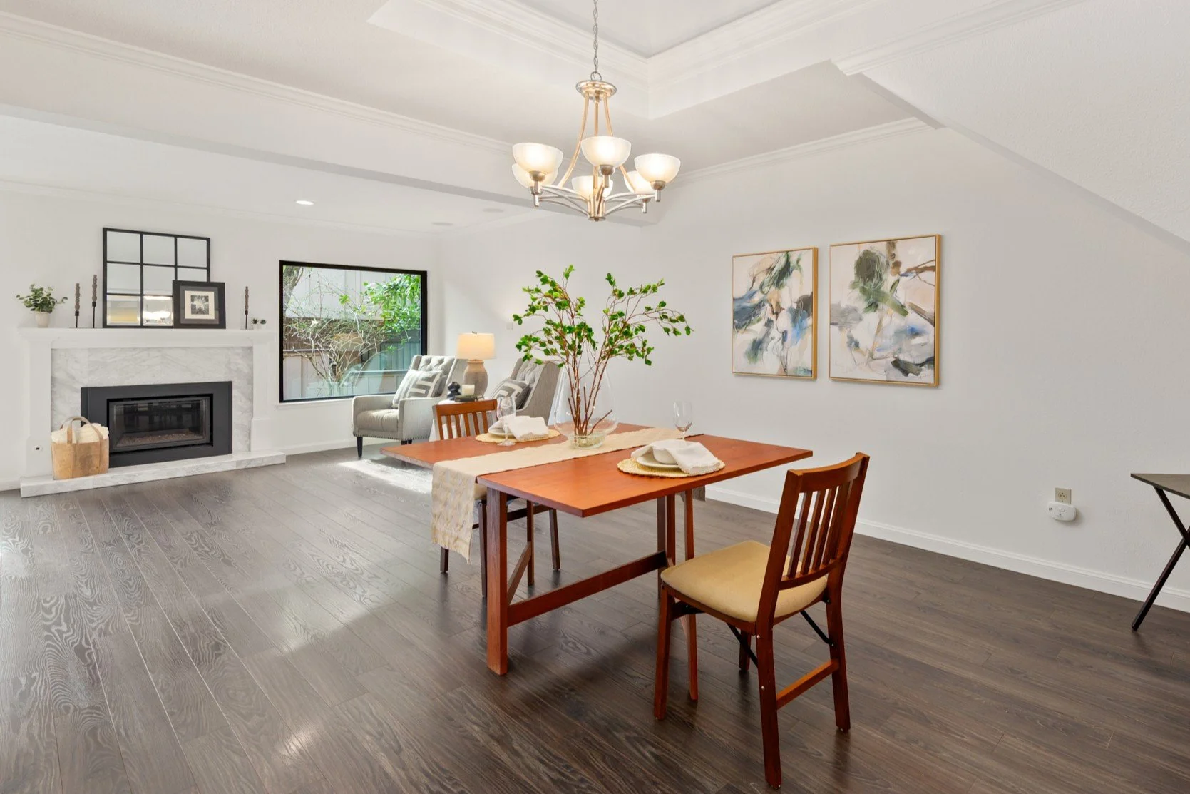 Open living and dining room with white walls, wooden flooring, fireplace, window, armchair, table with art, and a chandelier.