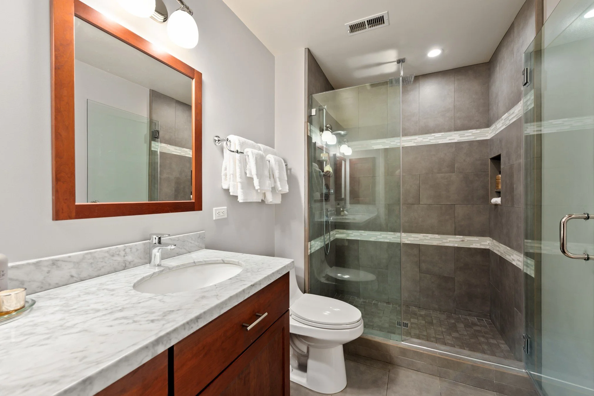 Modern bathroom with single sink vanity, marble countertop, mirror, wall-mounted lights, towel rack with white towels, toilet, and glass-enclosed shower with tiled walls and built-in shelf.