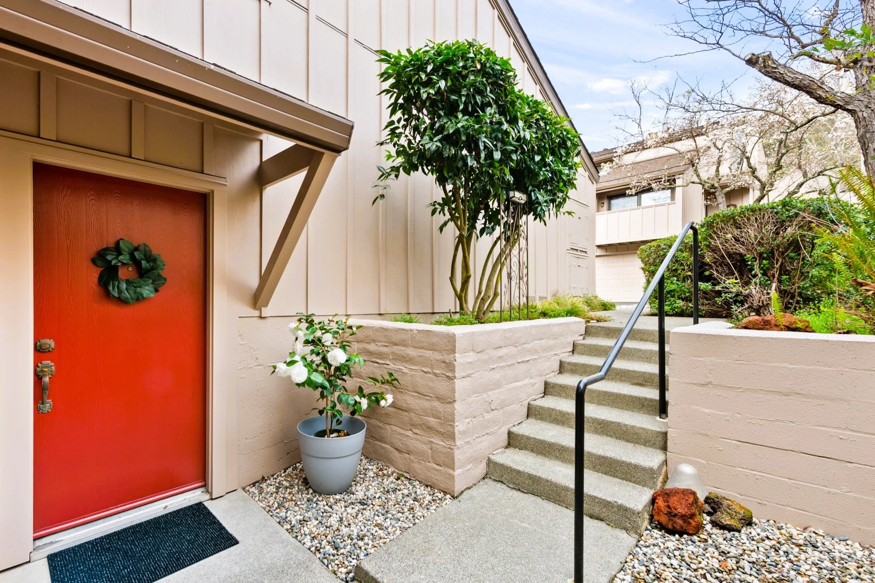 Exterior view of a house with a red door decorated with a wreath, a potted white flowering plant, concrete stairs with a black handrail, and a garden area with various bushes and trees.