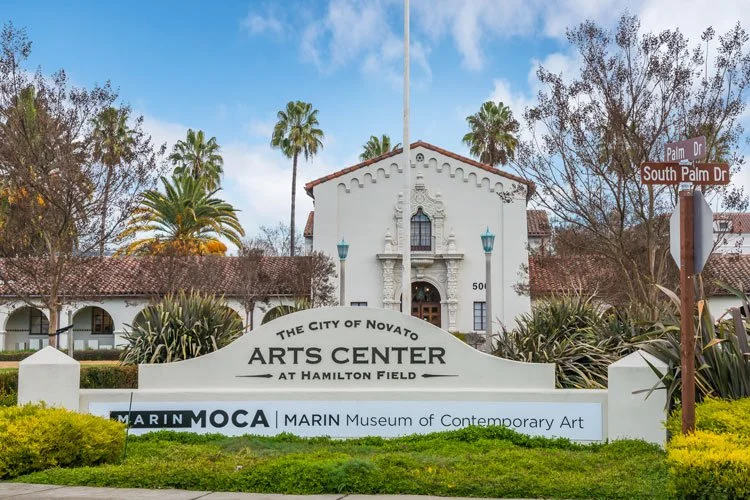 Sign for the City of Novato Arts Center at Hamilton Field, near a historic white building with palm trees and street signs for Palm Drive and South Palm Drive.