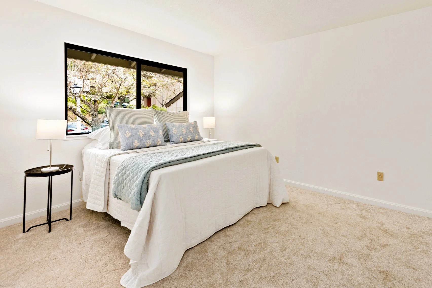 A clean, minimalist bedroom with a bed, side table, and lamps, next to a large window showing trees outside.