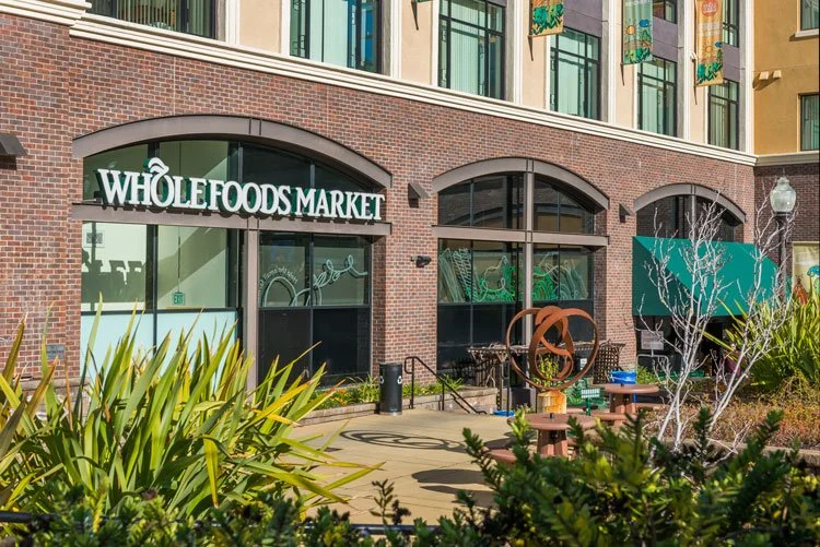 Exterior of a Whole Foods Market store with large glass windows, brick facade, and outdoor seating area surrounded by green bushes and plants.