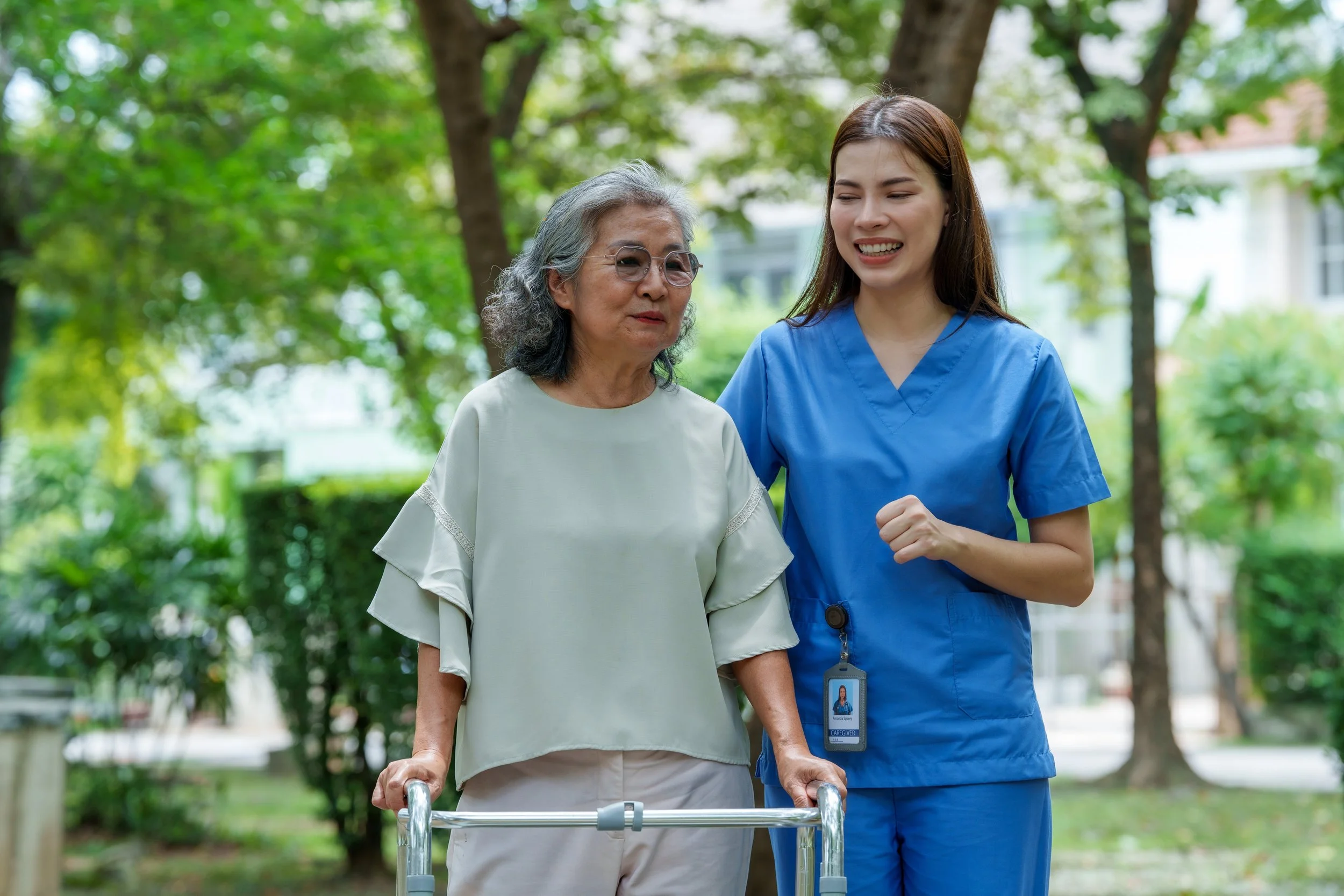 A young female healthcare worker in scrubs assisting an elderly woman with a walker outdoors in a park with green trees.
