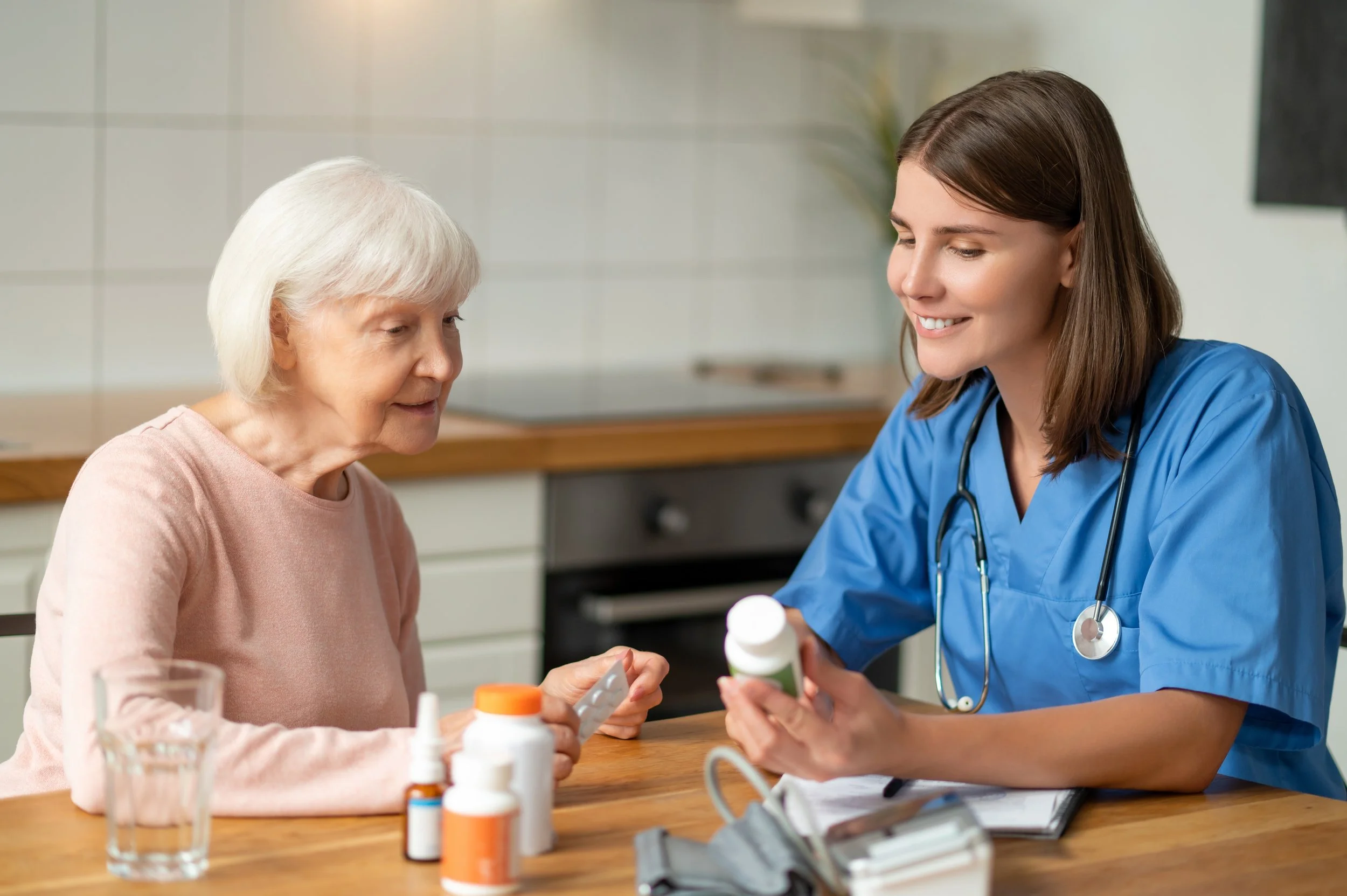 A young female nurse in blue scrubs and a stethoscope around her neck helping an elderly woman with medication at a kitchen table. The table has various pill bottles and a glass of water.