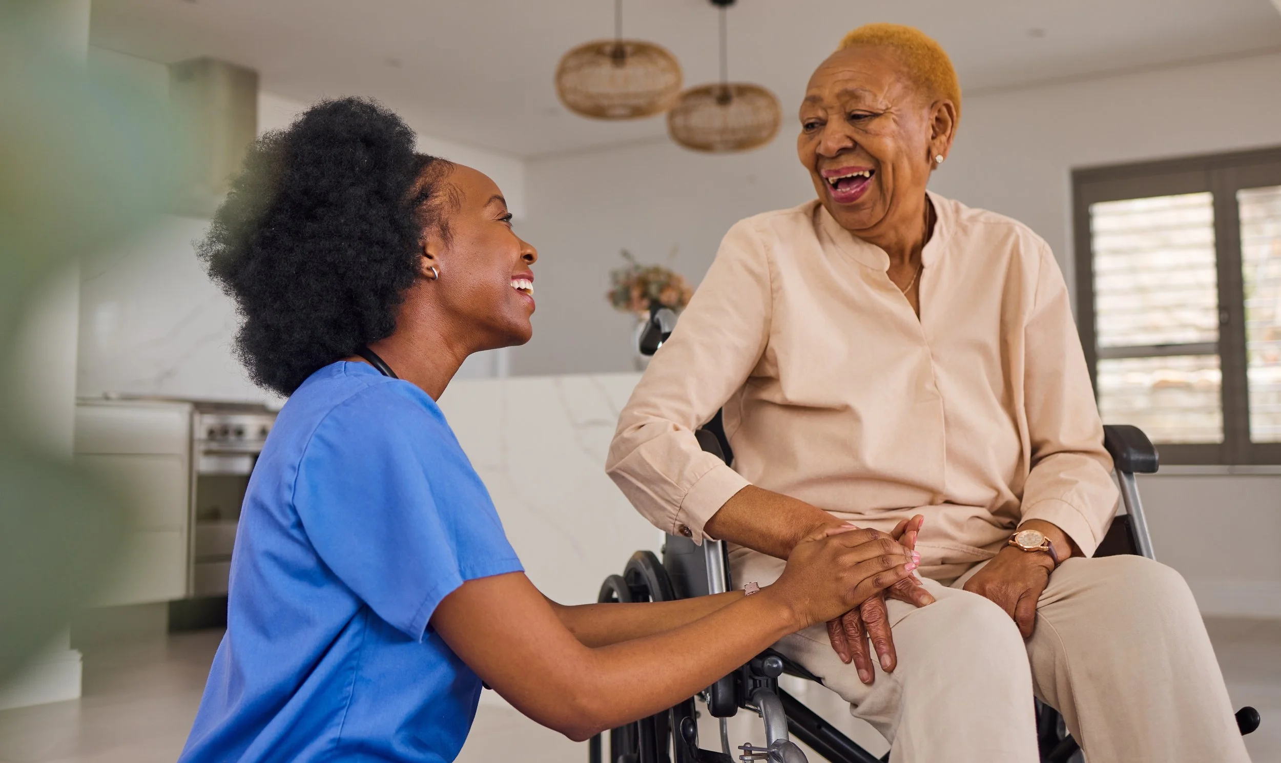 A young female healthcare worker in blue scrubs smiling and holding hands with an elderly woman in a beige blouse sitting in a wheelchair, both appearing happy and engaged in conversation indoors.