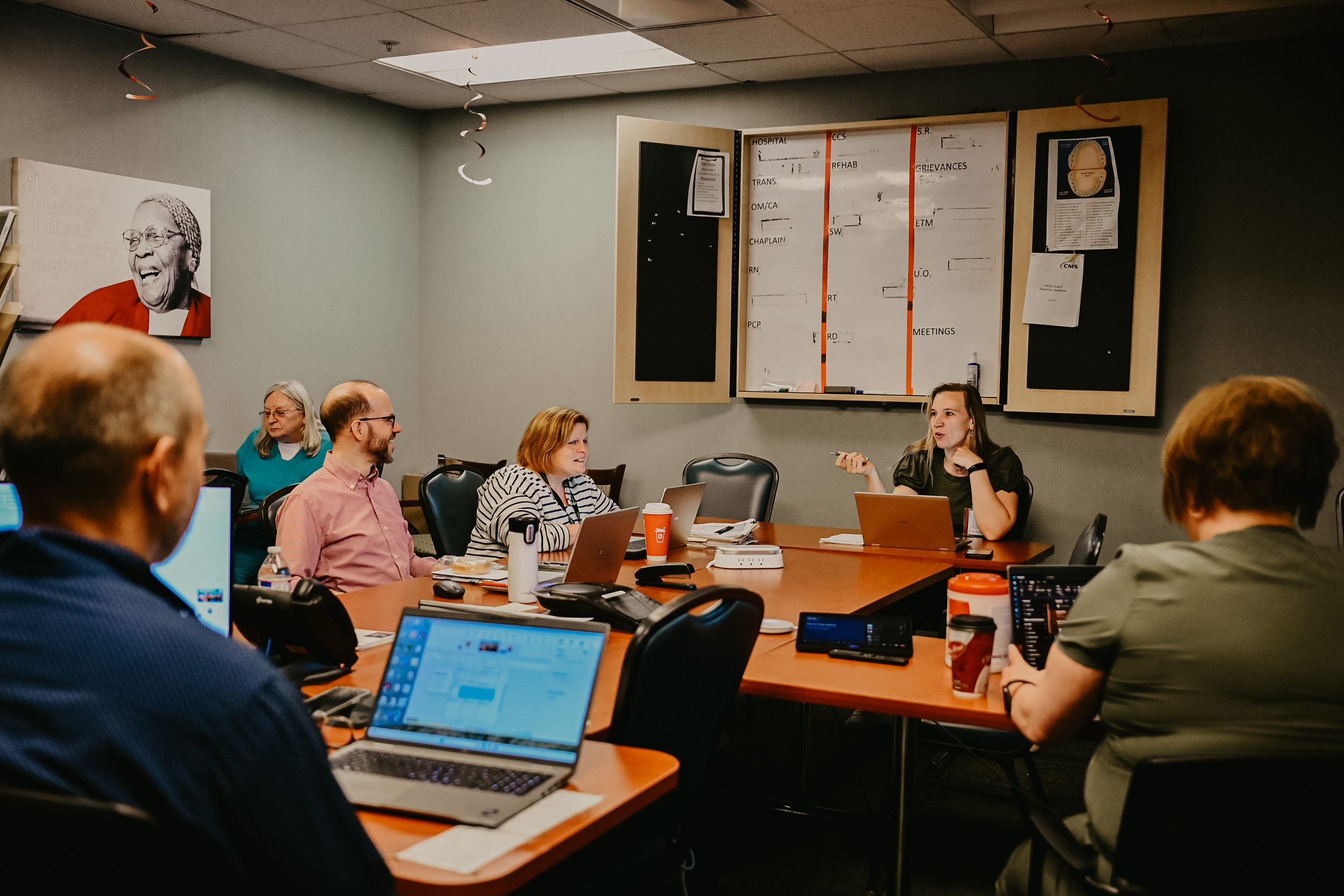 A group of people in a meeting room with laptops on a conference table, a whiteboard, and a large wall photo. LIFE Pittsburgh. Elderly