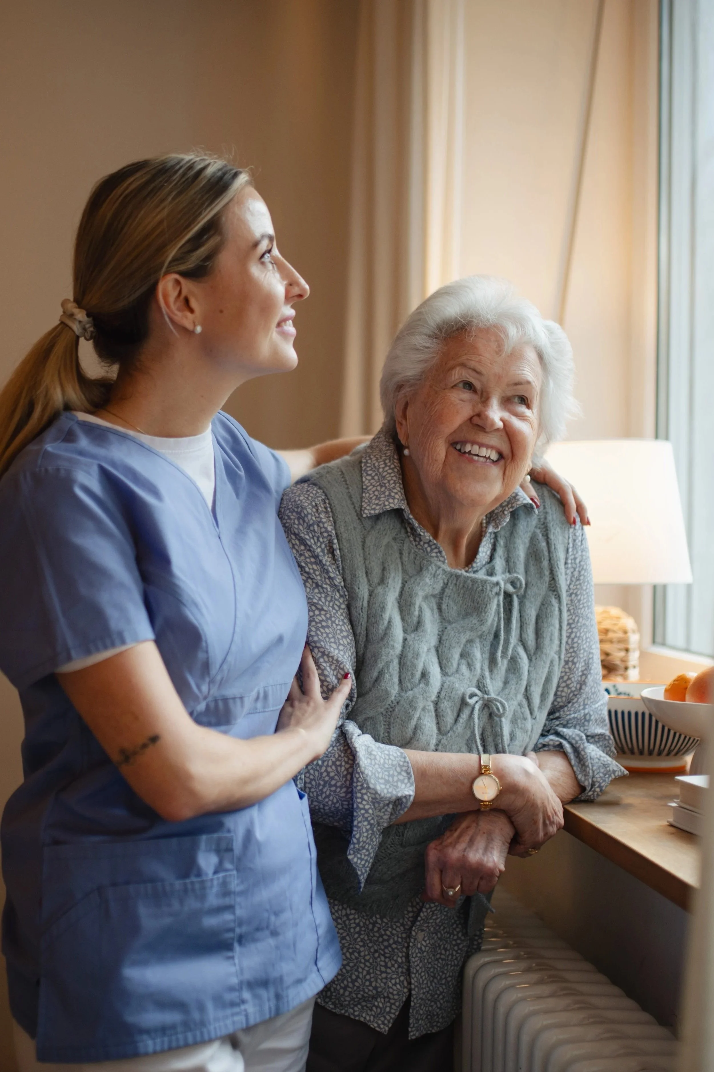 A young woman in medical scrubs stands beside an elderly woman, both smiling and looking out a window in a cozy room with warm lighting.
