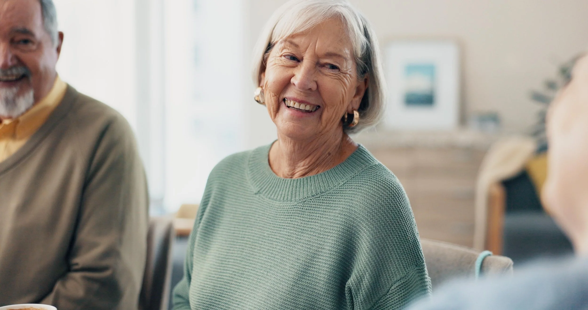 Smiling elderly woman with short gray hair wearing a light green sweater, sitting in a social setting with other elderly people visible.