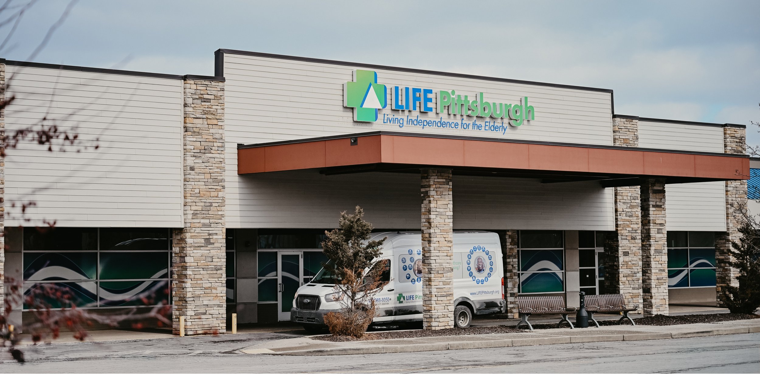 Exterior of a LIFE Pittsburgh Health and Wellness Center with LIFE Pittsburgh transportation vans parked outside, park benches and trees