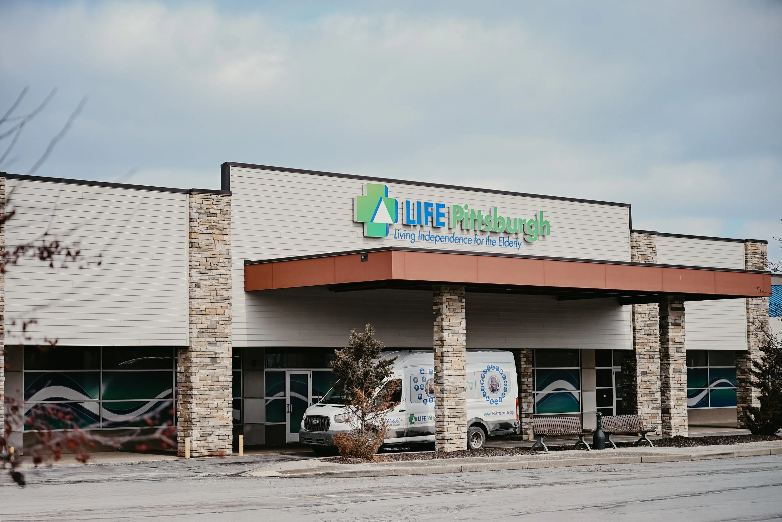 Front view of a building with a sign that reads 'LIFE Pittsburgh' and the tagline 'Living Independence for the Elderly.' There are parked vehicles, benches, trees, and cloudy skies.