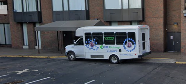 A white shuttle bus with decorations and text for Life Pittsburgh, parked on a city street in front of a brick building.