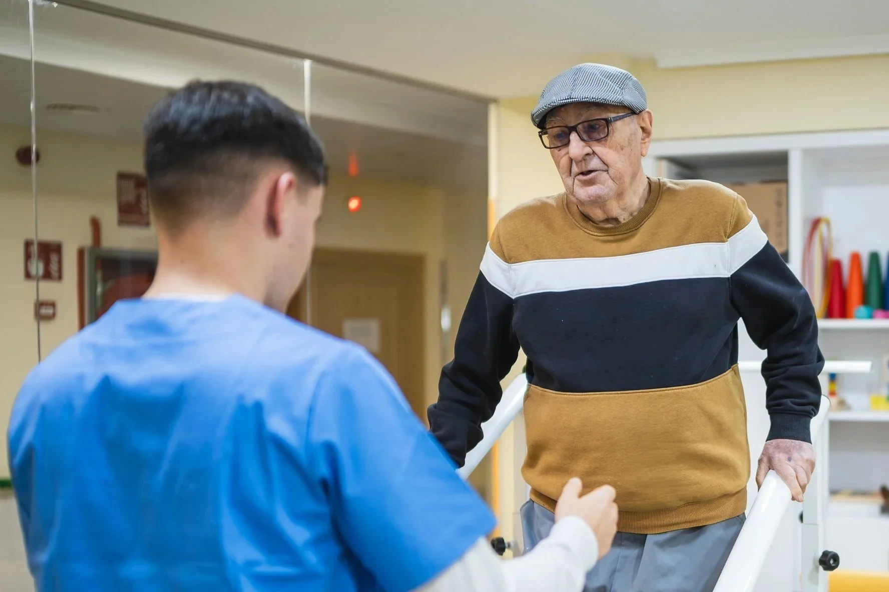 An elderly man with glasses and a gray cap is talking to a young man wearing a blue medical gown, in a hospital or medical facility.