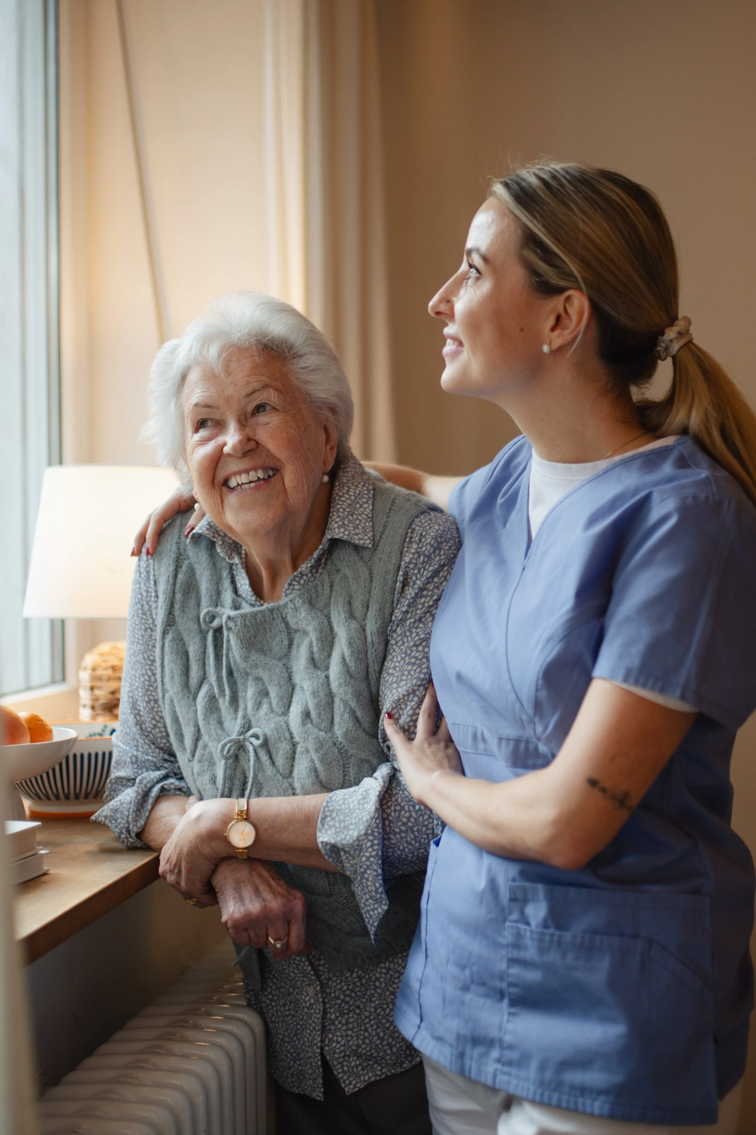 Nursing assistant smiling at an elderly woman in a cozy kitchen, both looking out the window.