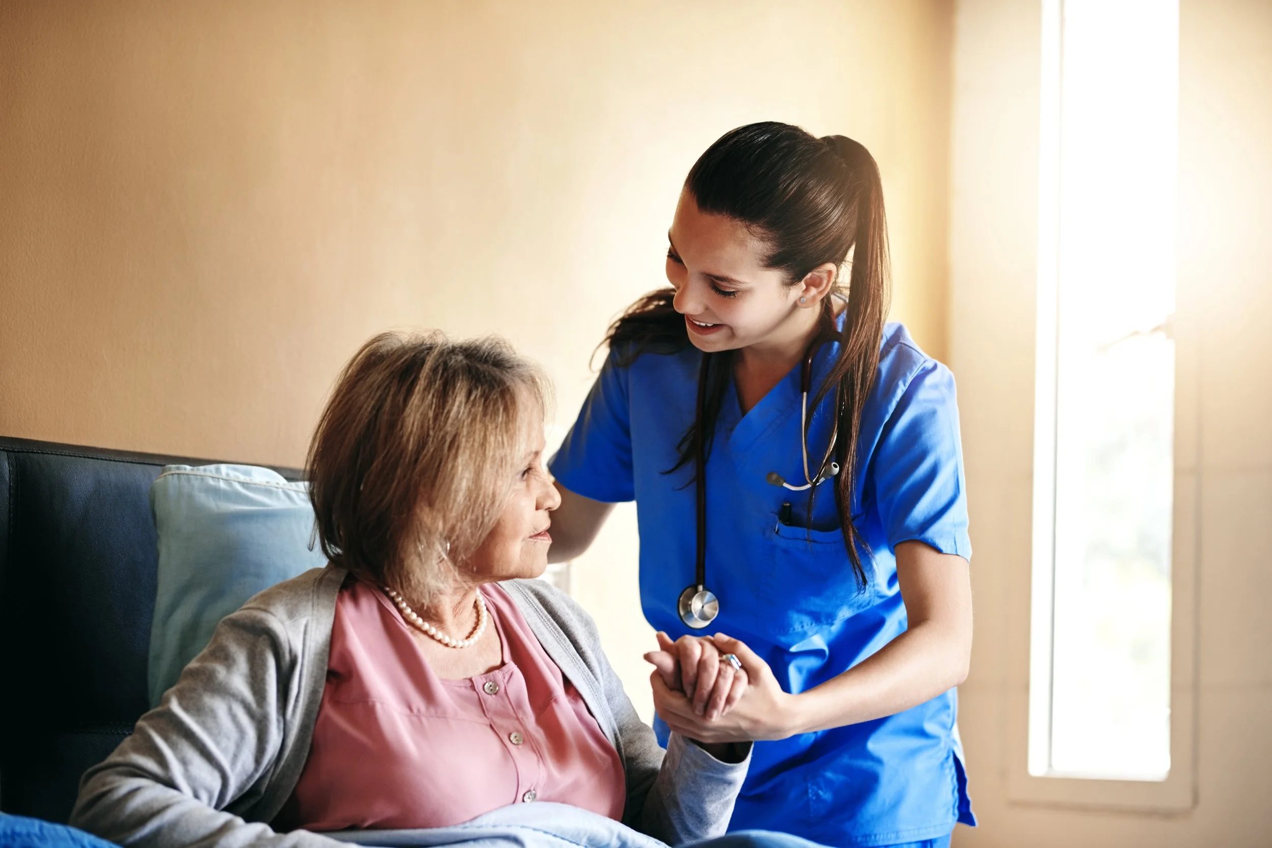 A female nurse or caregiver in blue scrubs with a stethoscope, smiling and holding hands with an elderly woman sitting in a hospital bed or chair.