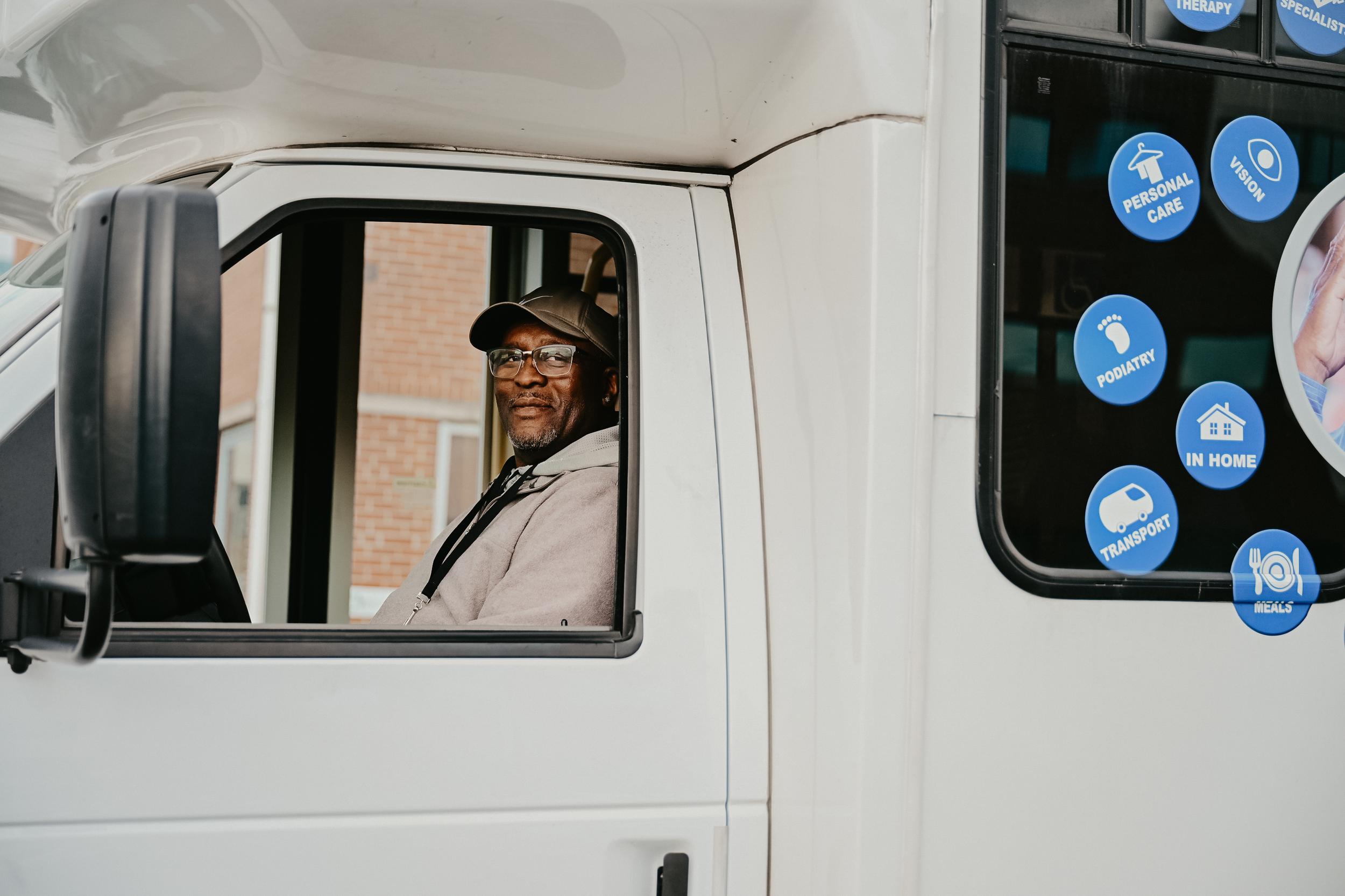A man sitting in the driver's seat of a white vehicle, smiling and looking out the window, with a brick building in the background and blue service department stickers on the window.