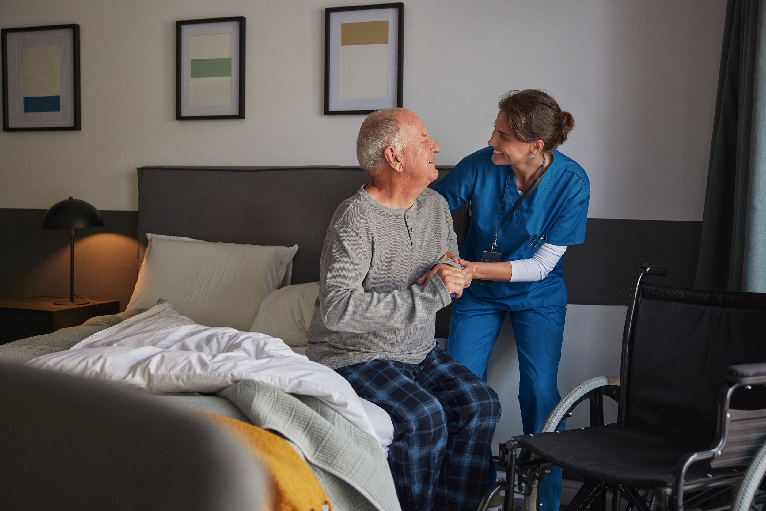 A nurse in blue scrubs smiling and holding hands with an elderly man sitting on a hospital bed, smiling back at her.