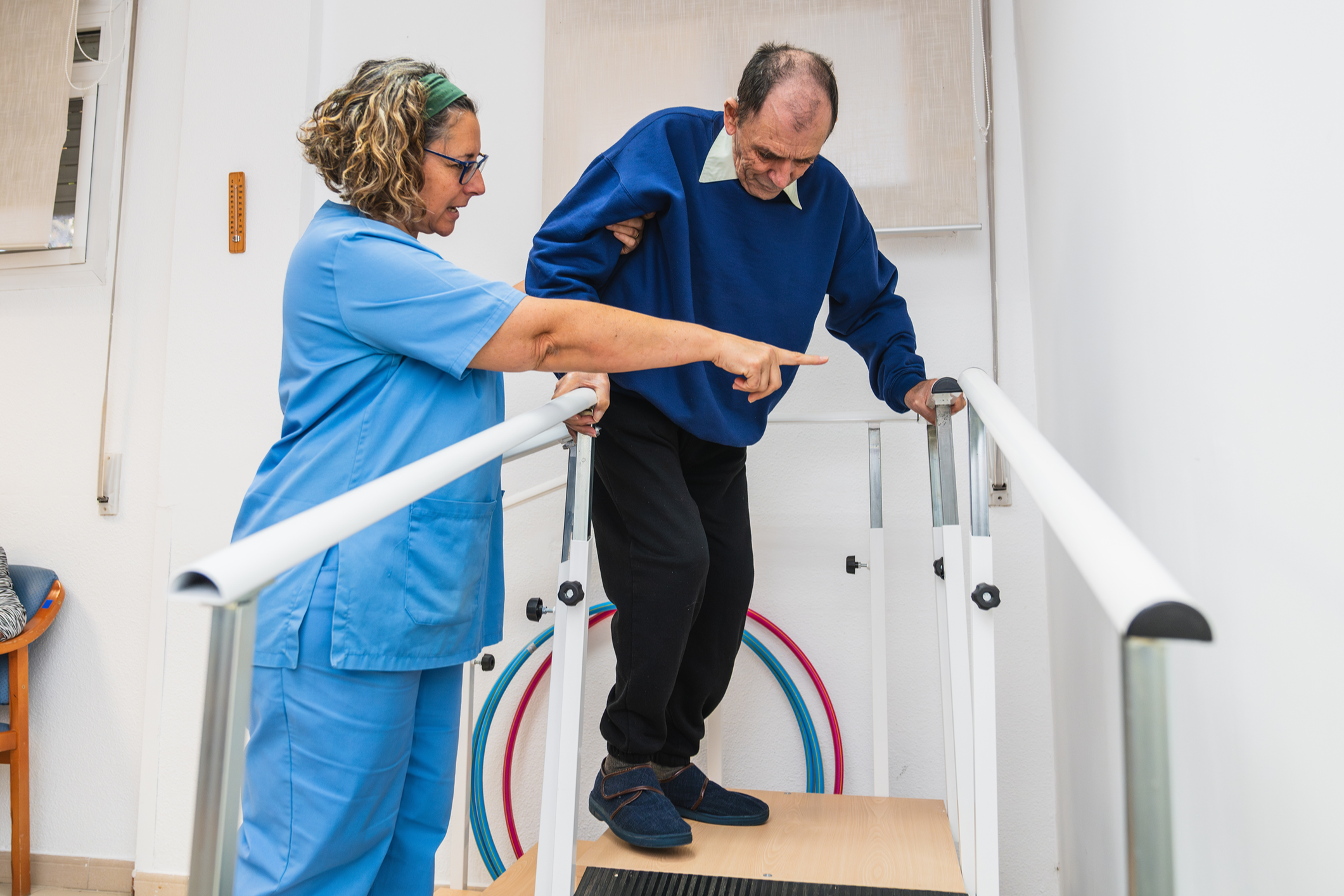 A caregiver assisting an elderly man in a blue sweater using a walker to ascend a set of stairs with support from a staff member in medical scrubs.