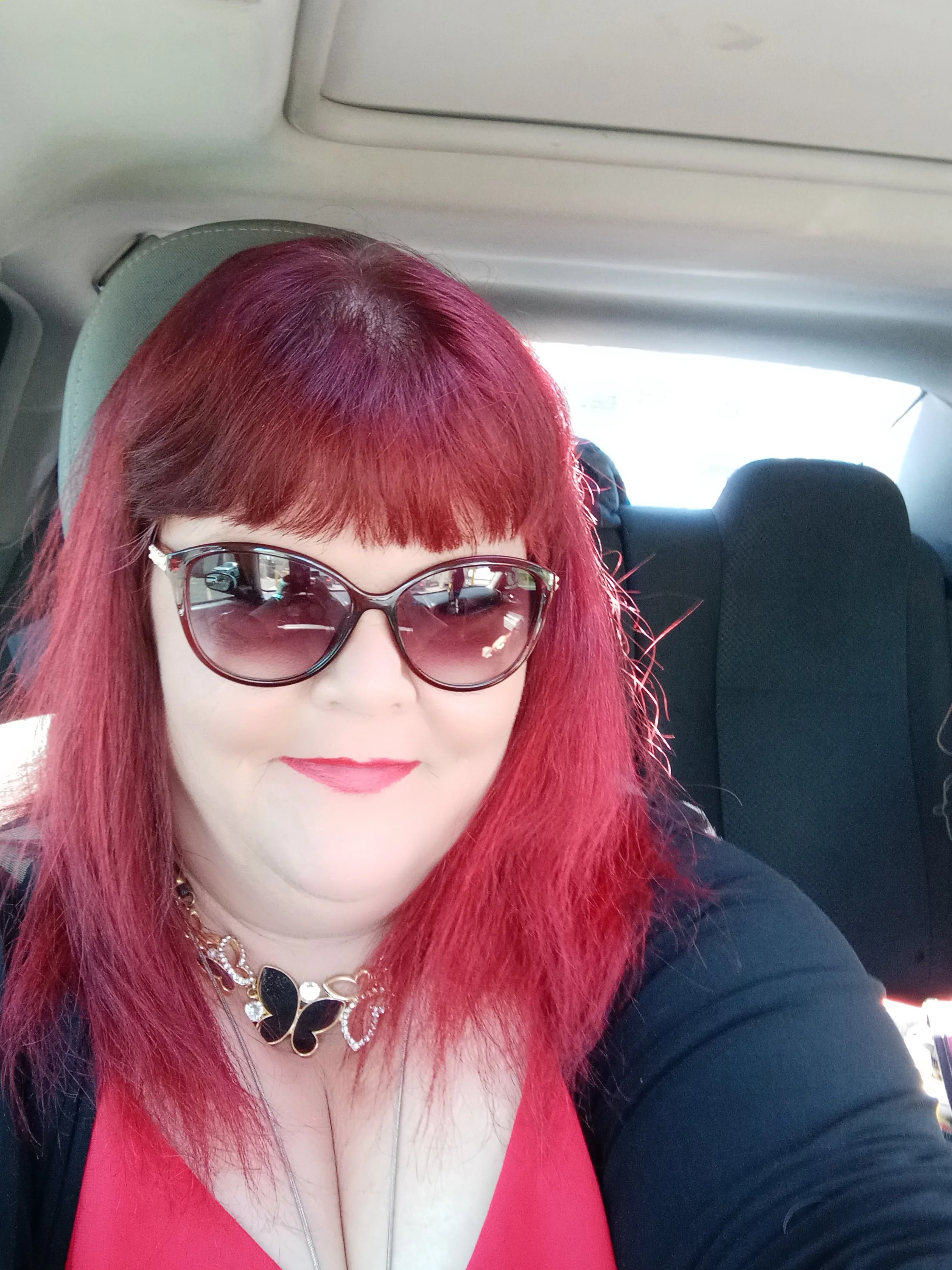 Woman with red hair, wearing sunglasses and a butterfly necklace, sitting in a car.