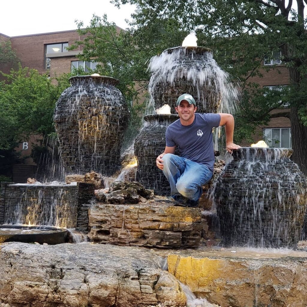A man kneeling on rocks in front of a multi-tiered outdoor fountain with cascading water, surrounded by green trees and a brick building in the background.