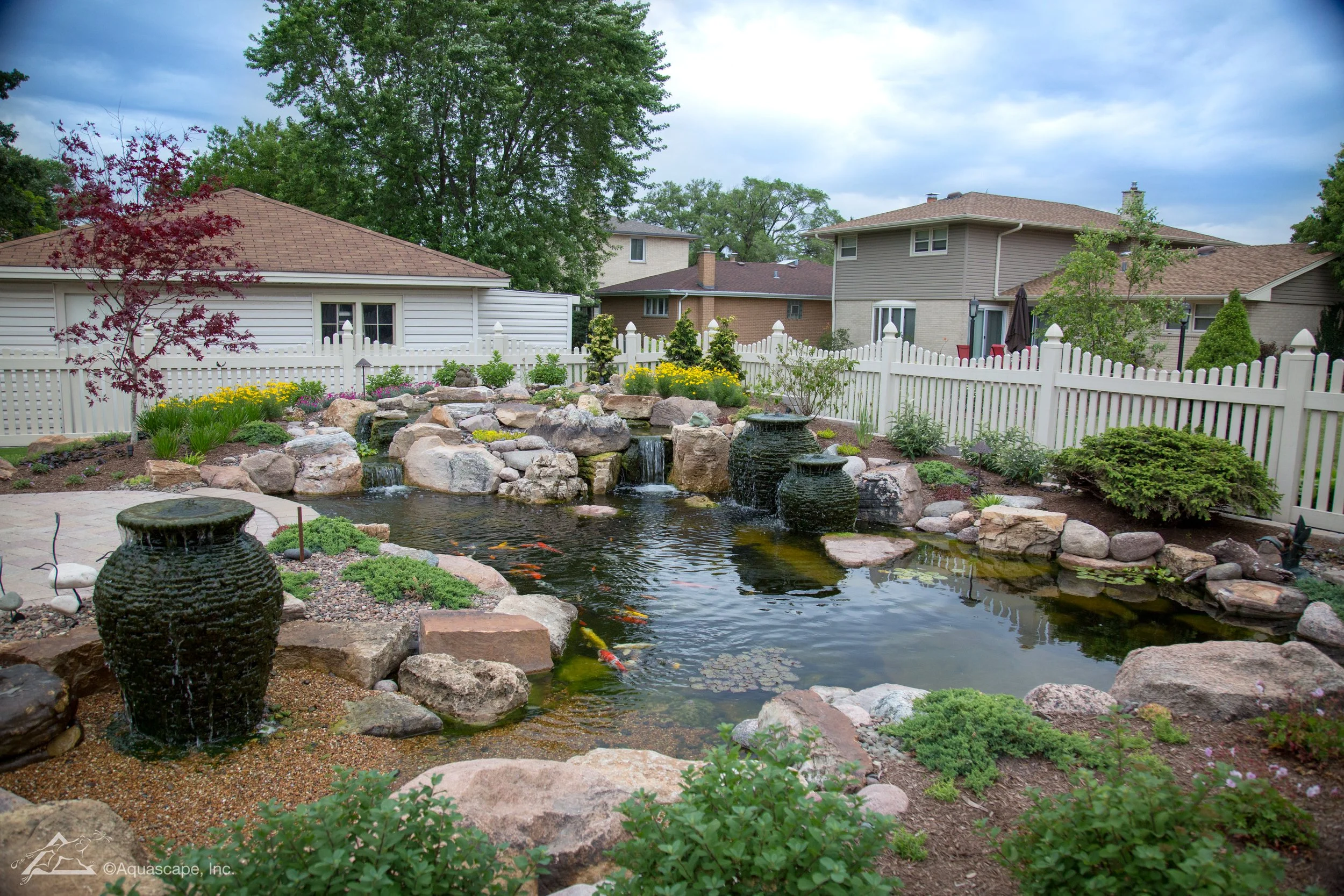 A backyard garden with a pond featuring waterfalls, surrounded by rocks, plants, and small trees. In the background, there are fenced-in yards and neighboring houses under a partly cloudy sky.
