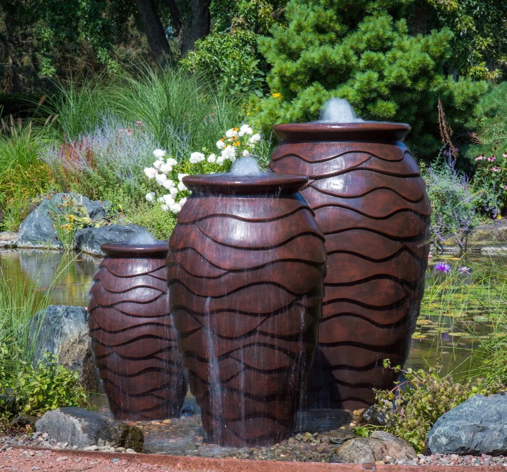 Three bronze-colored outdoor water fountains shaped like large jars with wave designs, with water flowing from the top, set in a lush garden with white, yellow, and purple flowers, green shrubs, and trees.