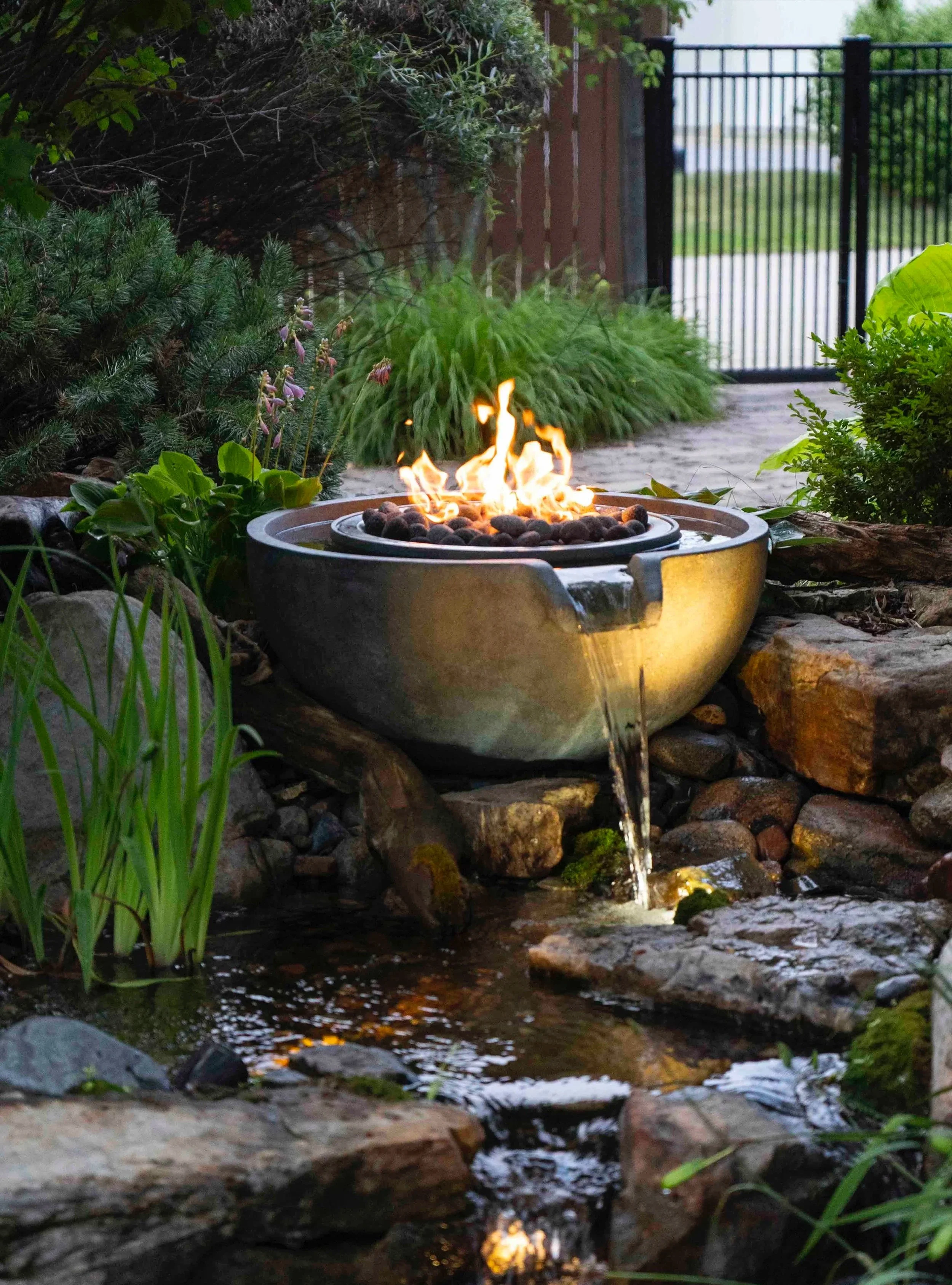 A small outdoor water fountain with a fire element on top, surrounded by rocks and lush greenery, with a black metal gate in the background.
