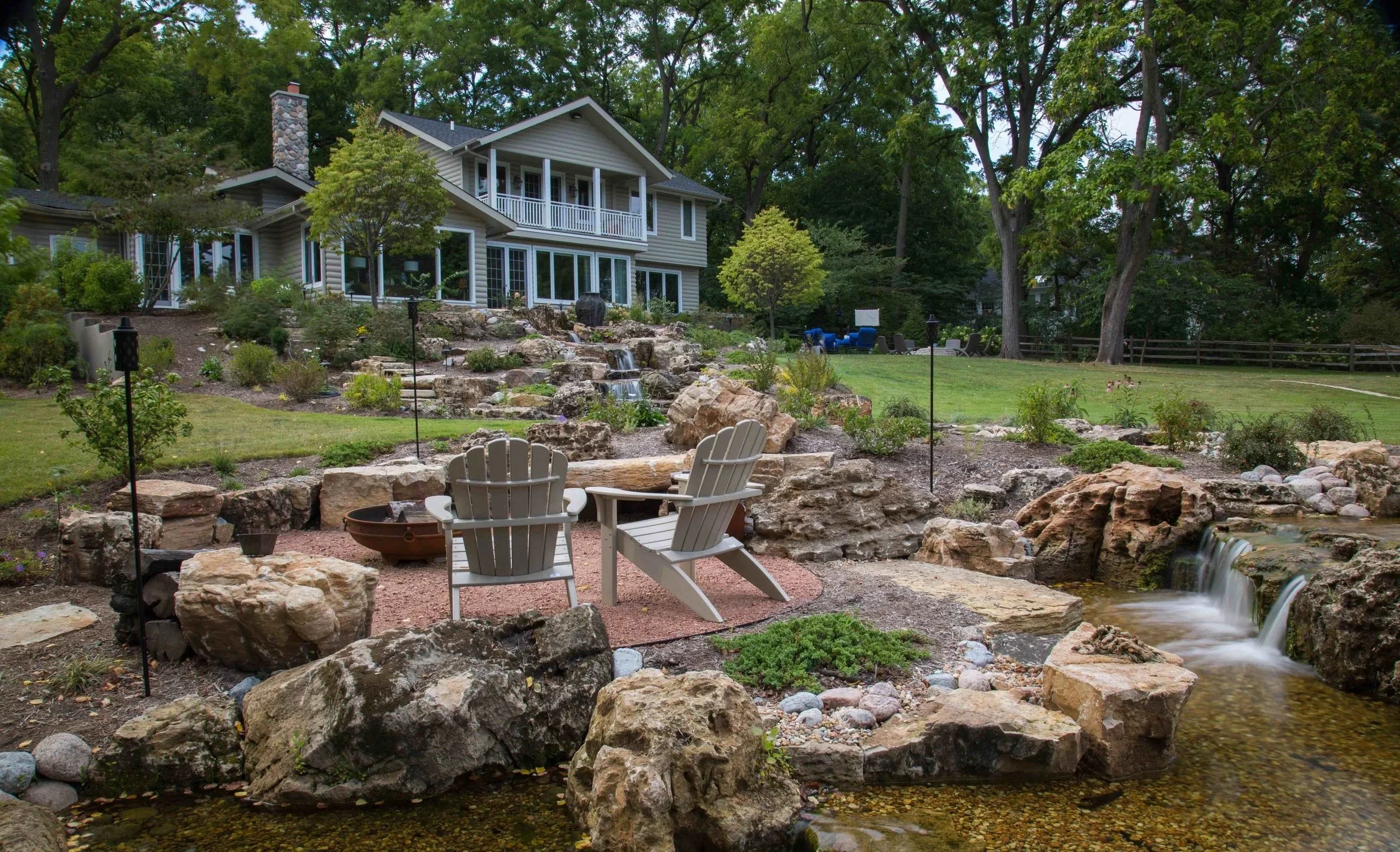 A landscaped backyard with a multi-story house, a small waterfall, a pond with rocks, Adirondack chairs, and tall trees.