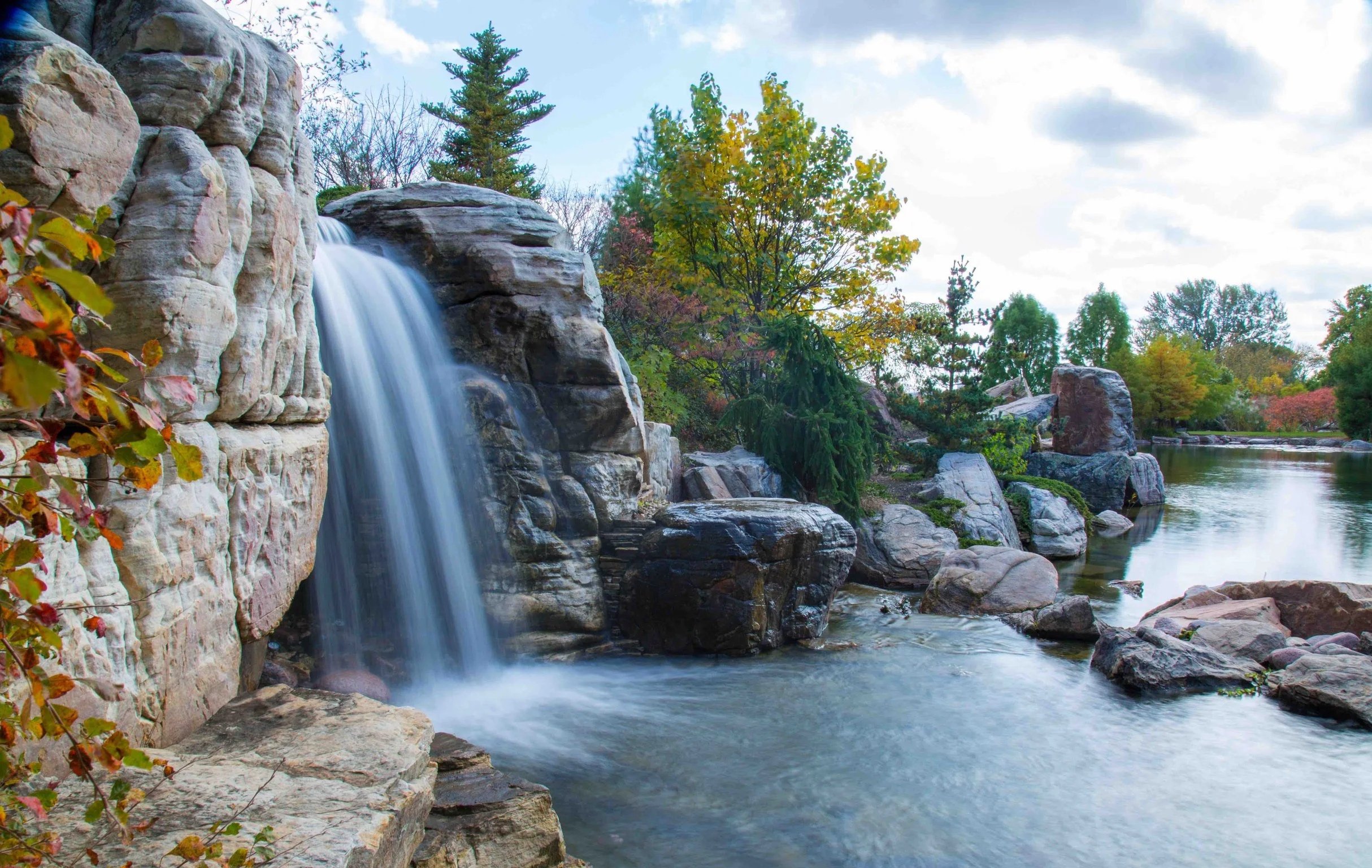 A landscape with a waterfall flowing over rocks into a calm river surrounded by trees with colorful autumn leaves, under a partly cloudy sky.